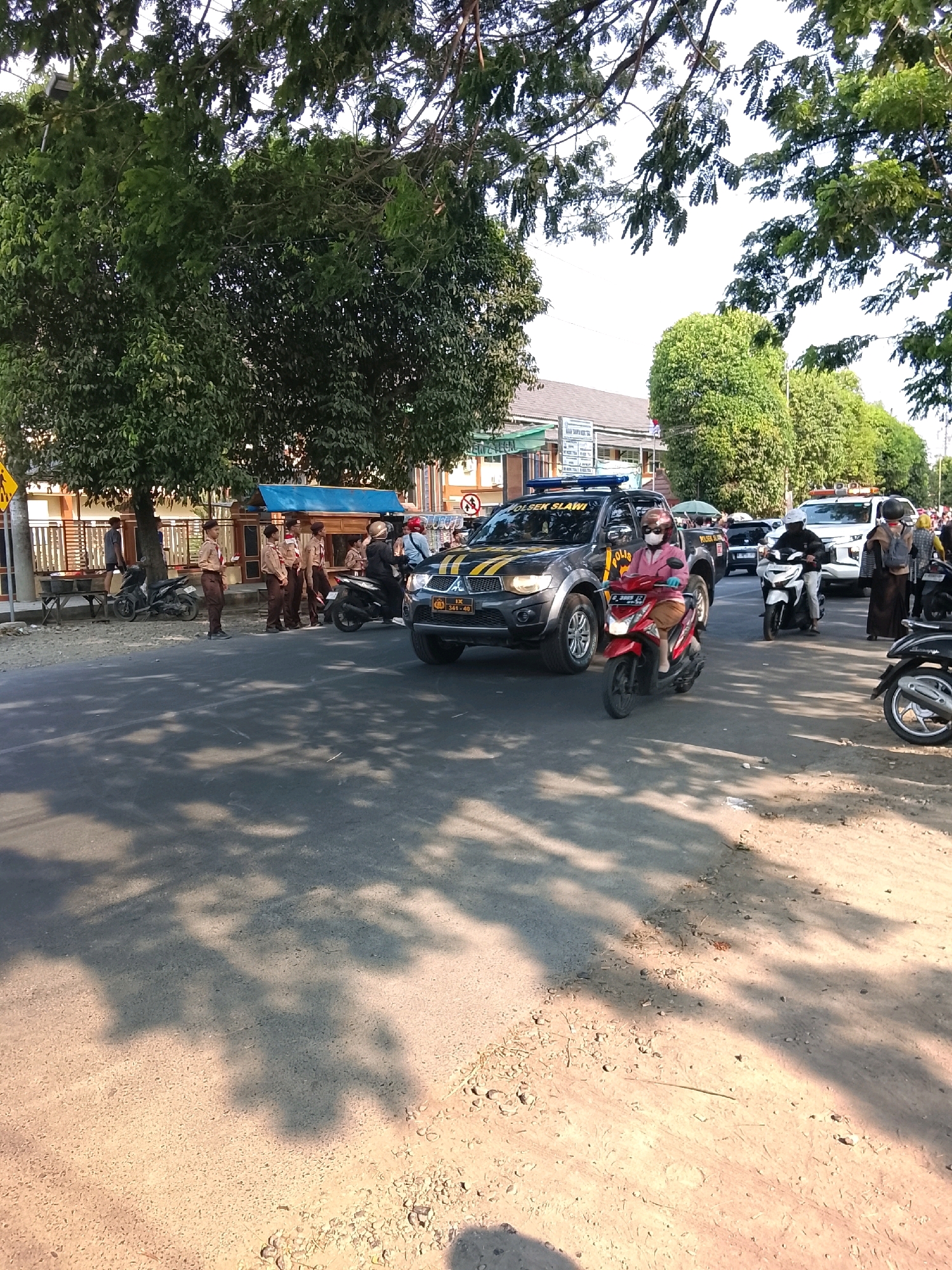 The image depicts a street scene in Slawi, Indonesia, with several people, vehicles, and buildings. In the foreground, a red scooter is ridden by a person wearing a pink shirt, white helmet, and face mask. To the right of the scooter, a black police SUV with yellow stripes and "POLSEK SLAWI" written on it is parked. A person in a dark jacket and helmet is on a white scooter behind the police SUV. Further to the right, a white police SUV is visible, with people standing and walking around it. In the middle ground, a group of individuals in brown scout uniforms are standing near a building with a blue awning. The street is paved and lined with trees, casting shadows on the ground. The environment appears to be an urban or semi-urban setting on a sunny day.