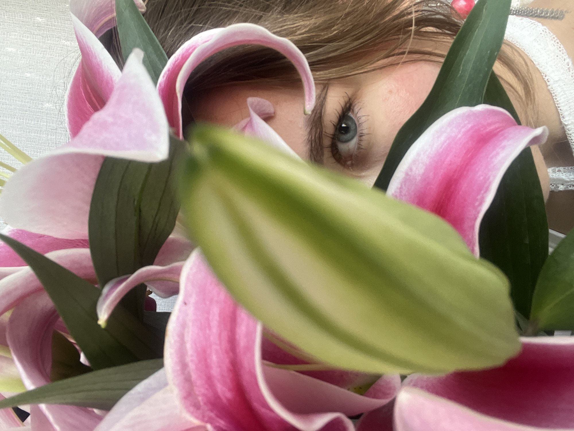 A close-up photograph shows a person's eye peeking through a cluster of pink lilies with green leaves and a green bud. The eye is blue with dark lashes. A portion of the person's forehead and hair are visible. Part of a white, lacy garment is visible on the right side of the frame. The photograph is taken indoors. The location and time are unknown.