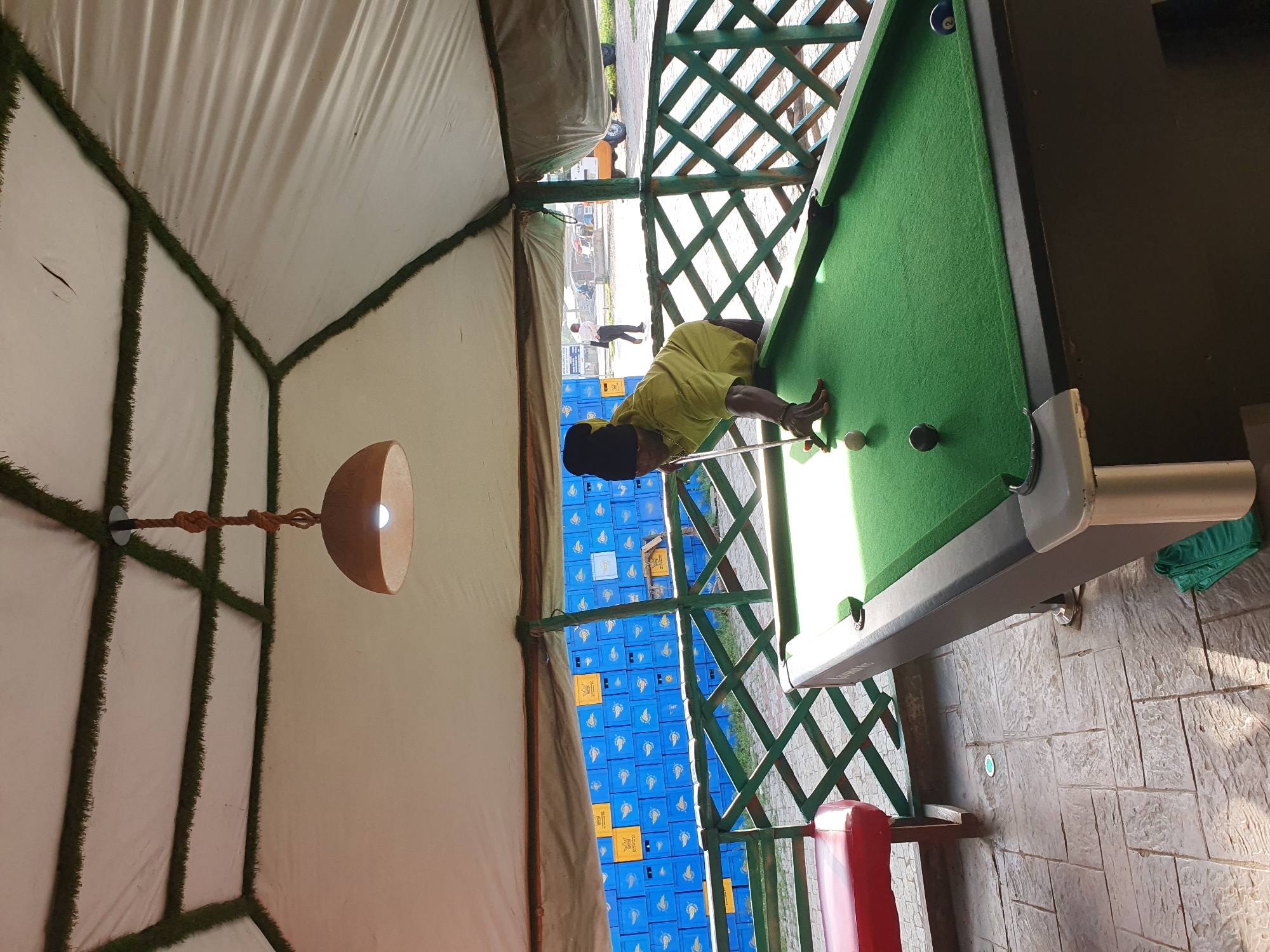 A person is playing pool under a tent. The tent has a white fabric roof with green artificial grass along the edges of the ceiling panels. A light fixture hangs from the center of the ceiling. The pool table is green with several balls on it. The person is wearing a yellow shirt and a black hat and is leaning over the table, aiming the cue stick. In the background, there are stacks of blue crates and a green lattice fence. The ground is paved with stone tiles. This scene appears to be an outdoor recreational area, possibly a bar or entertainment venue, in Lokoja, Nigeria.