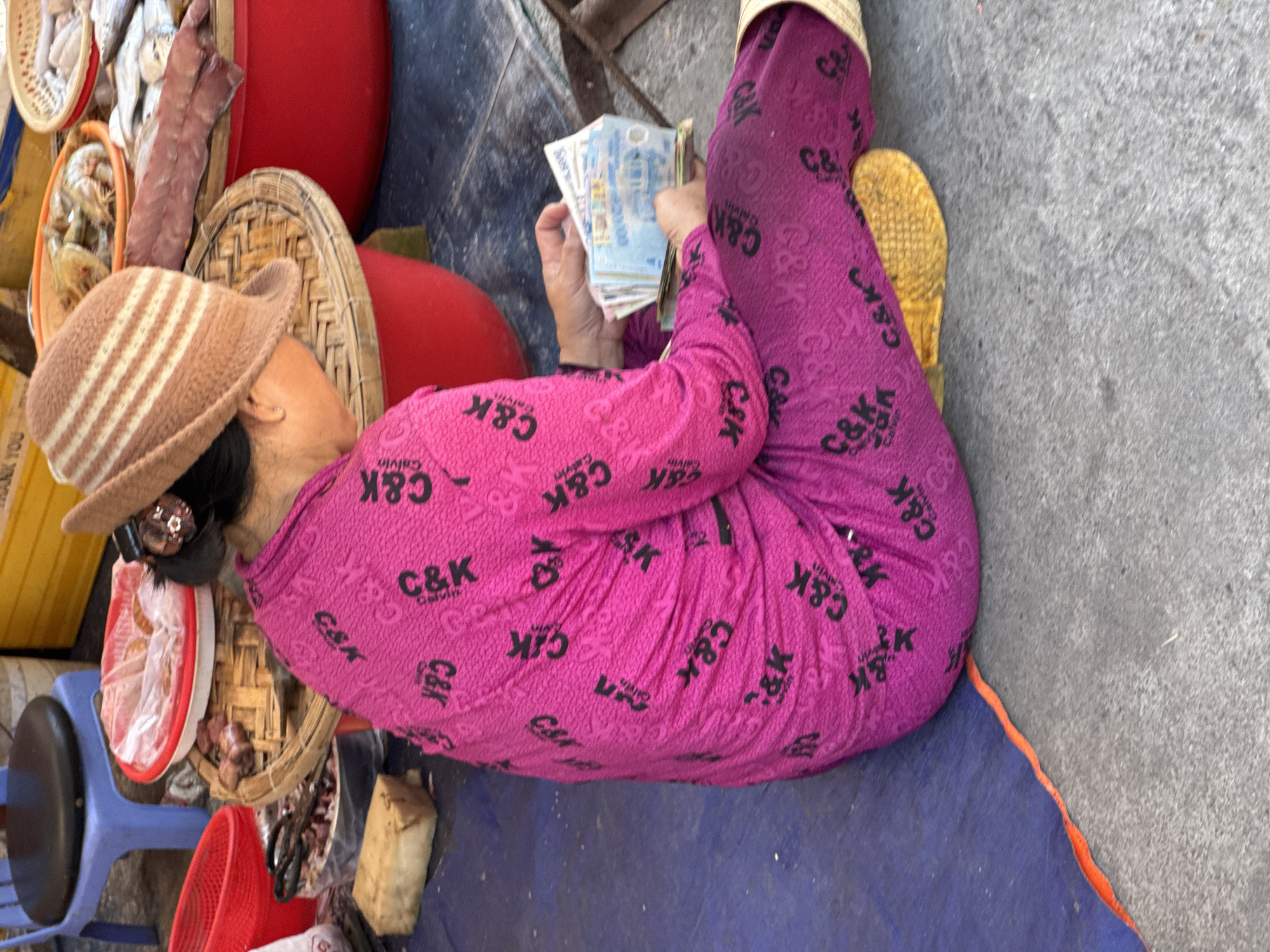 A woman, seen from behind and the side, sits on a blue tarp on a concrete surface, holding a wad of Vietnamese Dong currency in her hands. She wears a bright pink long-sleeved shirt and pants, both patterned with black "C&K" logos and "Calvin" text. A brown and white striped knit hat covers her head, and a small, decorative clip is visible in her hair. The background suggests a market scene: to her left, a blue stool and red plastic bins; behind her, a woven tray holding food items, and further back, a large red tub with fish. The lighting is natural, indicating an outdoor setting. The location is a market in Nha Trang, Vietnam.