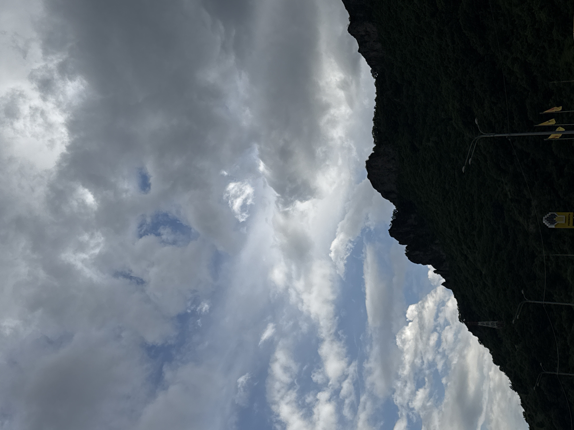 The image depicts a dramatic, cloud-filled sky over a densely forested mountainside. The sky is a mix of dark, heavy storm clouds and patches of lighter, scattered clouds, with glimpses of blue sky peeking through. The mountainside is dark green, appearing heavily treed and rugged, with rocky outcrops visible along the ridgeline. A transmission tower, with visible red and white sections, stands on the slope of the mountain. In the foreground, streetlights and several flags on poles are visible, suggesting a roadside or public area. The overall impression is of a vast, natural landscape under a dynamic sky. The location is the city of Karachayevsk, Russia.