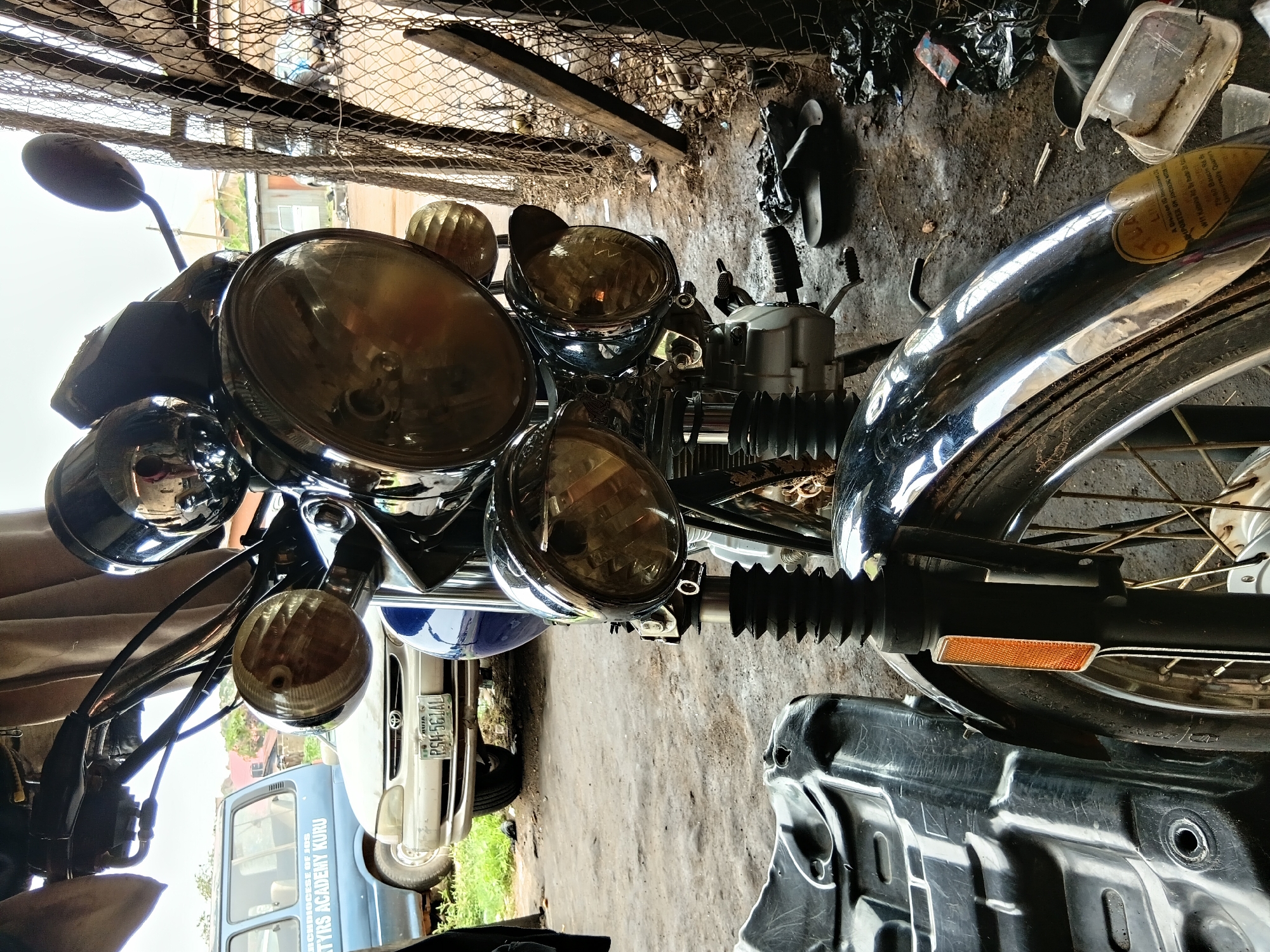 The image depicts a close-up view of a motorcycle parked outdoors. The motorcycle is a dark color, possibly black, with chrome accents on its headlights and other parts. It has multiple round headlights. In the background, a light-colored car and a blue van are partially visible. The van has text that reads "ARCHDIOCESE OF JOS MARTYRS ACADEMY KURU". A wire fence is visible to the right of the motorcycle, behind which is a cluttered area with debris and some discarded footwear. The ground is dirt or gravel. The scene appears to be in an open, possibly unkempt, area within the city of Jos, Nigeria, suggested by the text on the van.