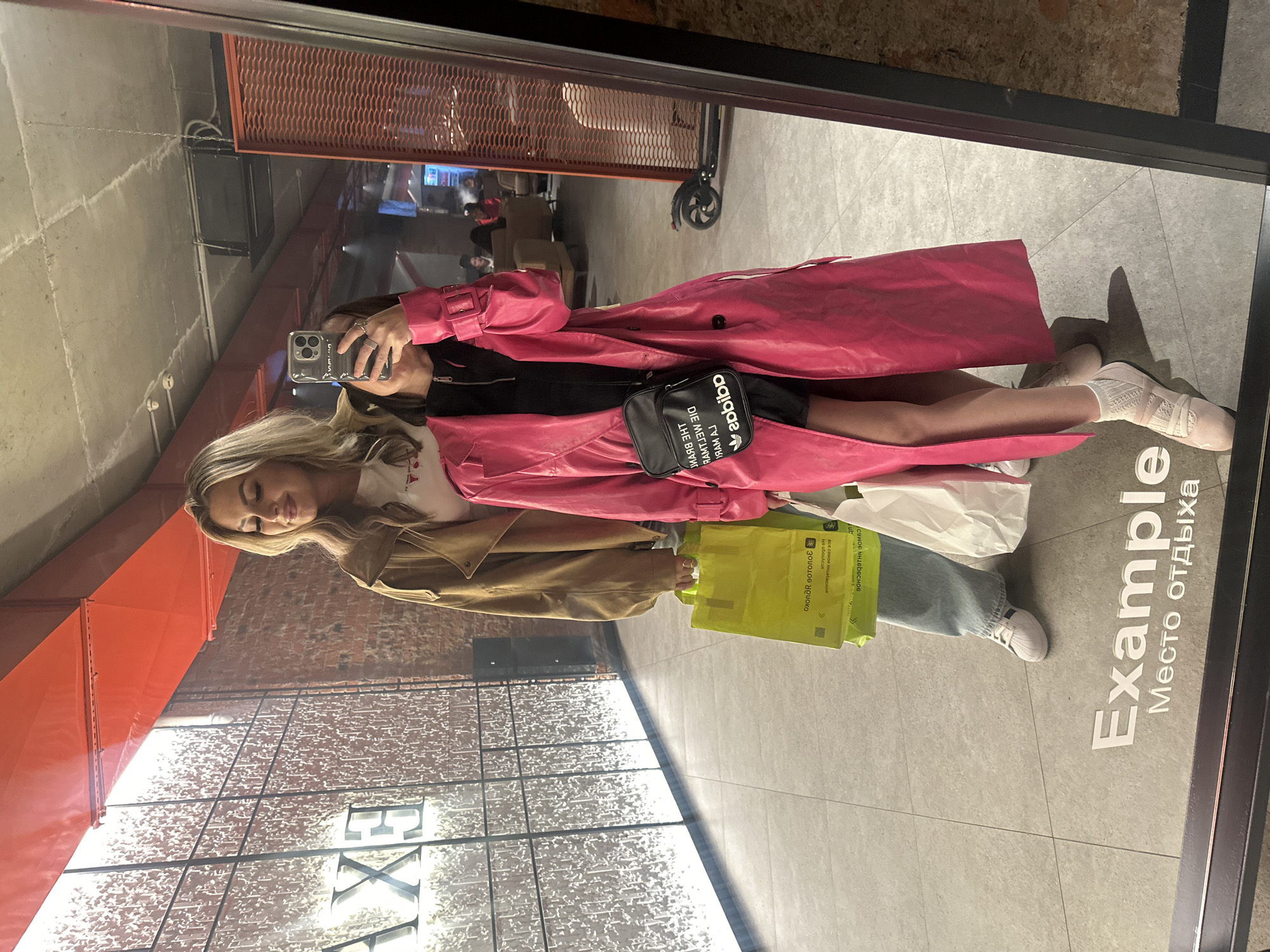 Two women are captured in a mirror selfie within a modern commercial or retail space in Kazan, Russia.
The woman on the left stands taller, possessing blonde, wavy, shoulder-length hair. She wears a light-colored t-shirt, a tan-brown oversized jacket, light blue jeans, and white Adidas Superstar sneakers. She holds multiple bright yellow shopping bags in her left hand, which display the mirrored text "ЗОЛОТОЕ ЯБЛОКО" (Zolotoye Yabloko - Golden Apple) and "МЕСТО ОДЫХА" (Mesto Otdykha - Place of Rest). Her head is slightly tilted, and her eyes are partially closed in a soft expression.
The woman on the right, taking the photo, has dark, short hair, partially obscured by her smartphone, which she holds in her right hand. She wears a vibrant, long pink trench coat over a black top with a zippered collar. A black Adidas crossbody bag with white "ADIDAS" lettering and "THE BRAND WITH THE 3 STRIPES" (reflected) hangs across her body. Her left hand is partially visible, possibly holding a white shopping bag. She wears light pink ballet flats over white socks. A silver-toned ring is visible on her right hand.
The setting features a light grey tiled floor, reflecting white text that reads "Example" and "Место отдыха" (Place of rest). Behind the women, the background includes a tall glass partition or wall with a textured pattern on the left, illuminated by a bright light source. Above this, prominent orange industrial-style ventilation ducts run across the ceiling. A section of exposed brick wall is visible to the right of the glass partition. In the deeper background, reflected through an opening, a dimly lit lounge area with upholstered seating and other individuals is visible. To the far right, an electric scooter is parked near a dark, corrugated metal panel. The ceiling appears to be concrete with exposed conduits and lighting fixtures.
