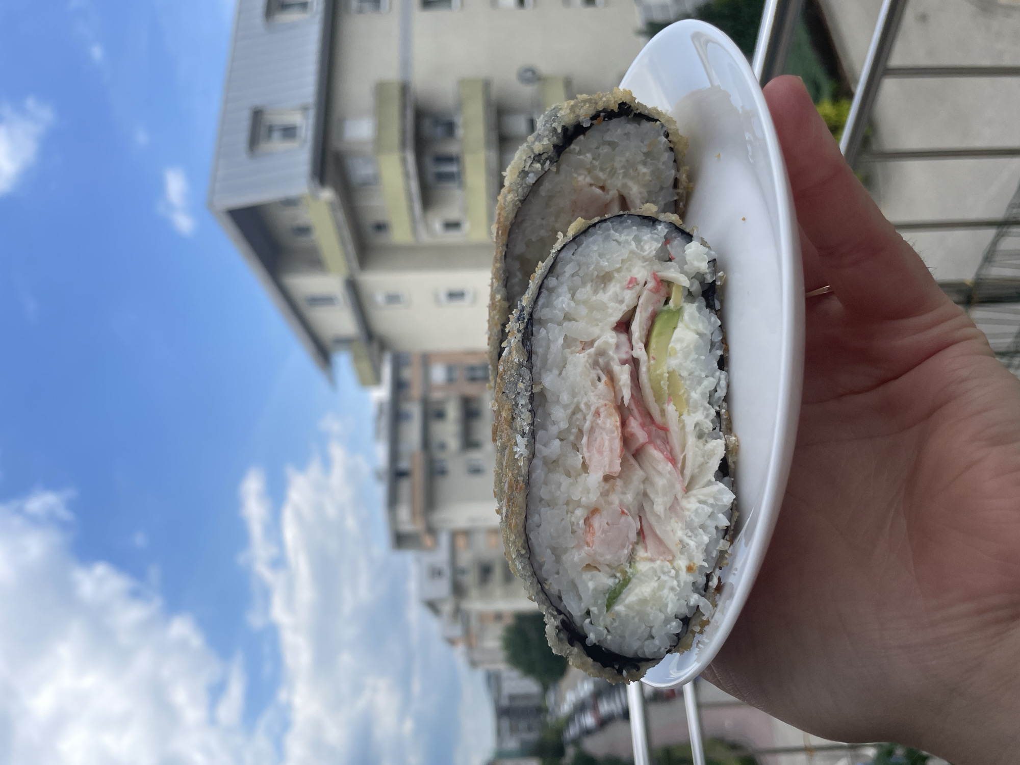 A person's hand holds a white plate with two halves of a fried sushi roll, cut to reveal the filling of rice, cream cheese, avocado, and imitation crab. The background shows a cloudy blue sky and the facades of apartment buildings.