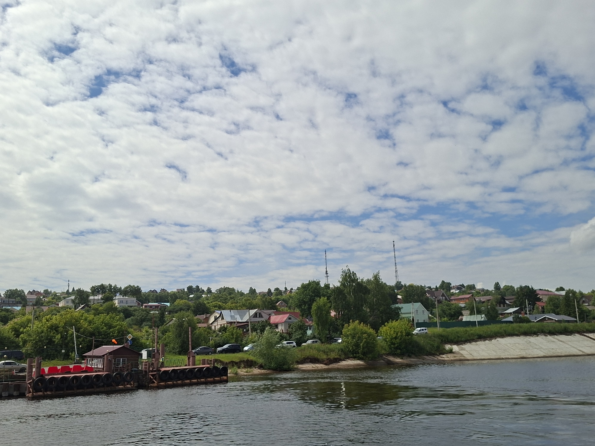 The image depicts a tranquil scene on a river in Verkhniy Uslon, Russia. In the foreground, a ferry dock is visible, constructed with dark, tire-like buffers along its side. A small, brown building with a red roof sits on the dock, and several rows of red seating are arranged behind it. The water in the river appears calm with slight ripples.
The middle ground showcases a verdant hillside densely covered with trees and houses. The architecture of the houses varies, with some featuring red roofs and others in lighter colors. A few cars are parked along the roadside that winds through the settlement. In the distance, two tall radio towers are prominently visible on the horizon. The sky is partly cloudy, with a mix of white cumulus clouds scattered across a blue expanse. The overall impression is one of a peaceful, semi-rural setting.