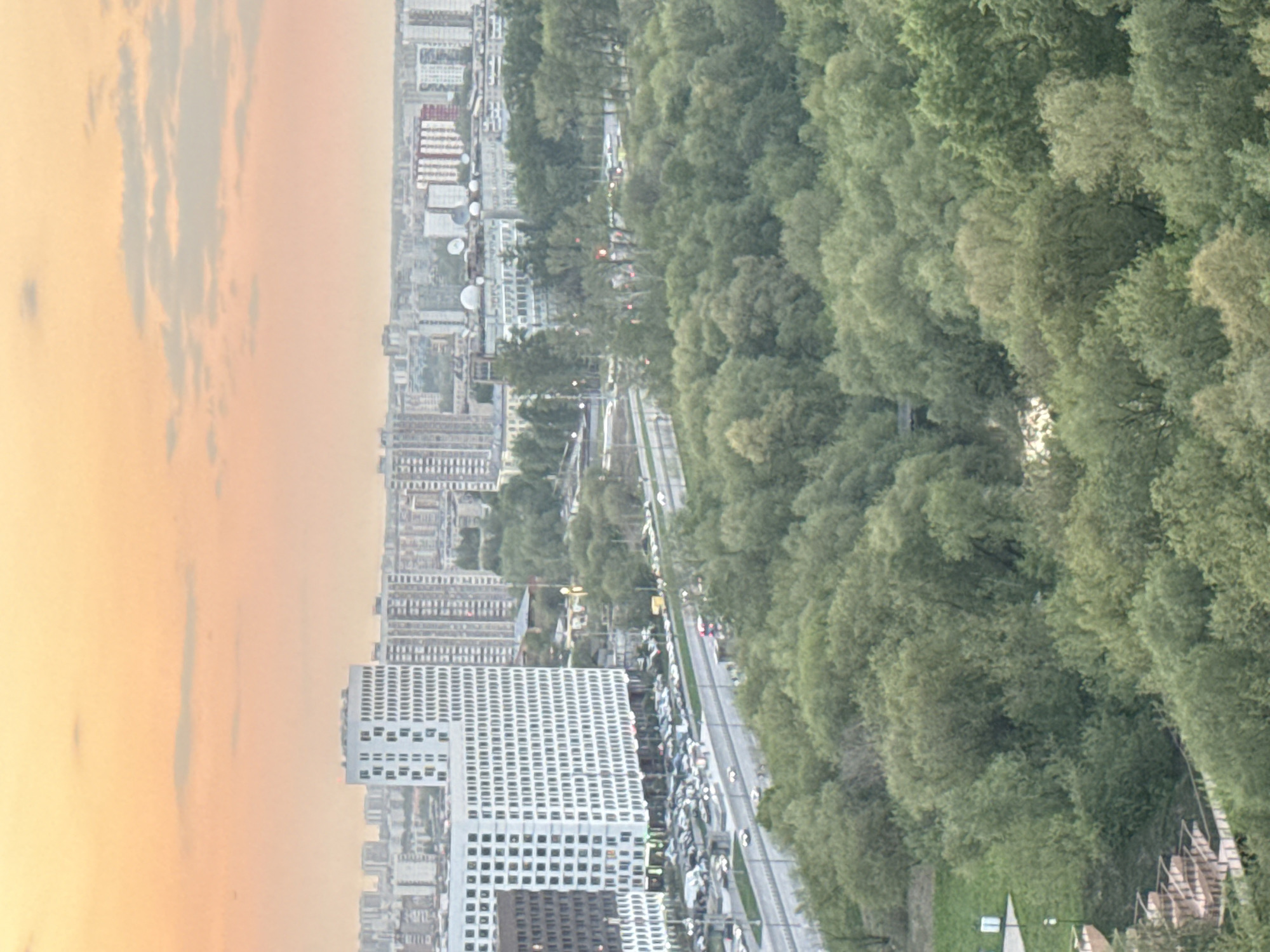 The image depicts a cityscape at sunset, with a dense cluster of modern high-rise buildings dominating the foreground. These structures feature a grid-like pattern of windows, suggesting residential or office use. A major road runs alongside the buildings, densely packed with vehicular traffic. Adjacent to the road, a large expanse of lush green trees extends into the distance, forming a park or forested area. The sky is filled with muted orange and pink hues, indicating the time of day. The location appears to be an urban area with a significant green space bordering it.