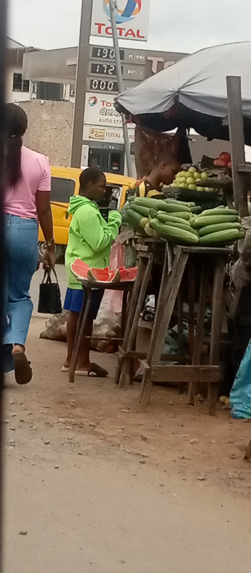The image captures a street scene in Onitsha, Nigeria, likely a market area given the presence of vendors and merchandise. In the foreground, a woman in a bright green hoodie and blue shorts stands behind a display of sliced watermelons. She appears to be interacting with a customer, although the customer is not fully visible. To her left, another person, mostly out of frame, is wearing a pink shirt and blue jeans, carrying a black bag.
Behind the fruit stall, a young person, possibly a boy, is seen tending to a variety of produce, including cucumbers and small yellow fruits, likely lemons or limes. The stall itself is constructed from rough wooden planks, covered by a light-colored umbrella.
In the background, a yellow truck is parked. Further back, a Total gas station sign with price displays for petrol and diesel is visible, indicating the presence of a commercial area. The ground is a mix of dirt and scattered debris. The overall impression is one of a busy, everyday commercial transaction taking place in an open-air environment.
