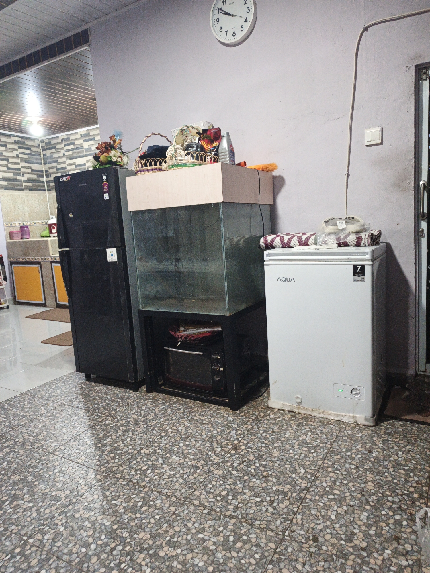 In Kelapapati, Indonesia, a cluttered indoor scene depicts a domestic living space. A large black refrigerator stands to the left, adorned with stickers. Next to it is a glass aquarium on a black stand, housing an unseen interior and what appears to be an oven below. To the right, a white chest freezer with the "AQUA" logo is topped with rolled-up mats and plastic-wrapped items. Above the appliances, a large round clock with black numerals hangs on a pale purple wall. A light-colored kitchen backsplash with a tiled pattern is visible in the background. The floor is covered in a mosaic of small, pebble-like tiles in shades of grey, white, and beige. The overall impression is one of everyday domesticity, with items arranged for practical use.