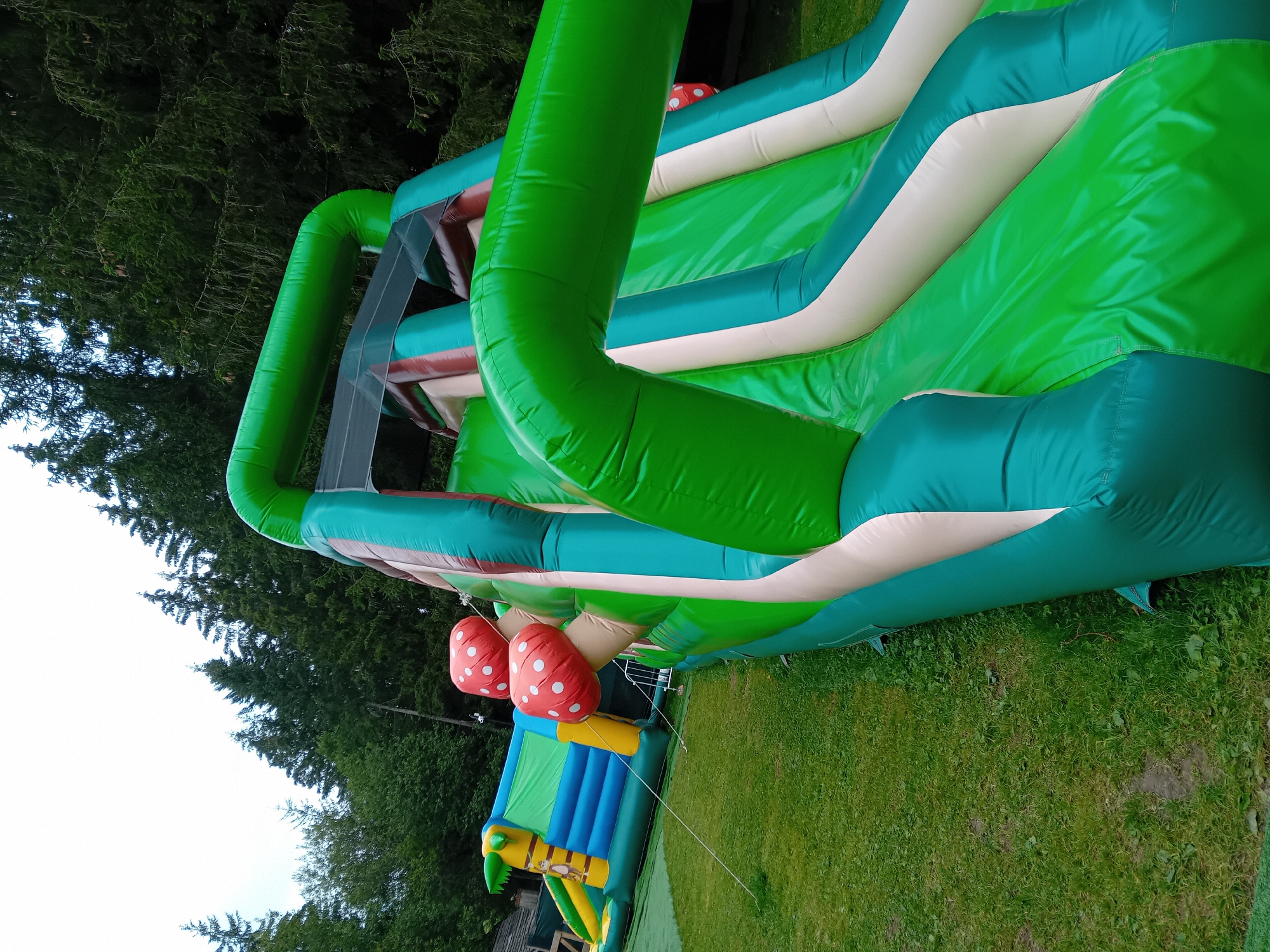 The image shows a vibrant, inflatable slide complex set up on a grassy area in Zuberec, Slovakia. The slide, primarily in shades of green and white, features a prominent arched structure and decorative red mushroom-shaped elements. In the background, a second, smaller inflatable attraction with a jungle theme is visible, featuring blue, yellow, and green colors and a cartoon monkey graphic. The area is surrounded by a dense forest of evergreen trees, indicative of a natural, possibly mountainous, setting. The sky is overcast. The scene appears to be an outdoor recreational area, likely a playground or amusement park, designed for children's entertainment. There are no people visible in the image.