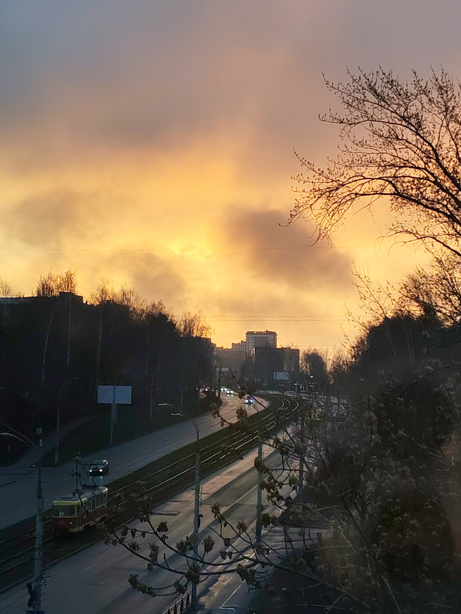 The image depicts a cityscape in Izhevsk, Russia, under a vibrant, orange-hued sky suggesting either sunrise or sunset. A four-lane road stretches into the distance, curving slightly. A tram runs parallel to the road. Light poles line the road, casting reflections on the wet pavement. Trees line both sides of the road. Buildings are visible in the distance. Bare tree branches frame the view in the foreground. A passing car in the road and a tram are in motion.