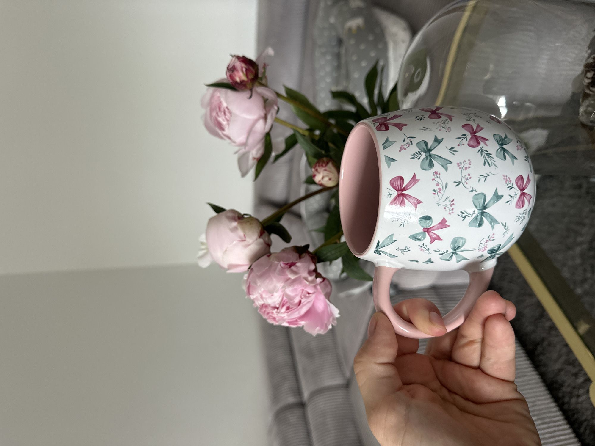 A close-up, eye-level, indoor shot focuses on a hand holding a white mug with a pink handle, adorned with a pattern of pink and teal bows and small sprigs of flowers. The mug is positioned in the foreground. In the mid-ground, a bouquet of three light pink peonies with green leaves is displayed in a clear vase, partially visible behind the mug. The background reveals a section of a grey, tufted sofa and a light grey wall, suggesting an interior setting. The overall lighting is soft and natural. There is no indication of the city of Kutenholz, Germany, in the image itself.
