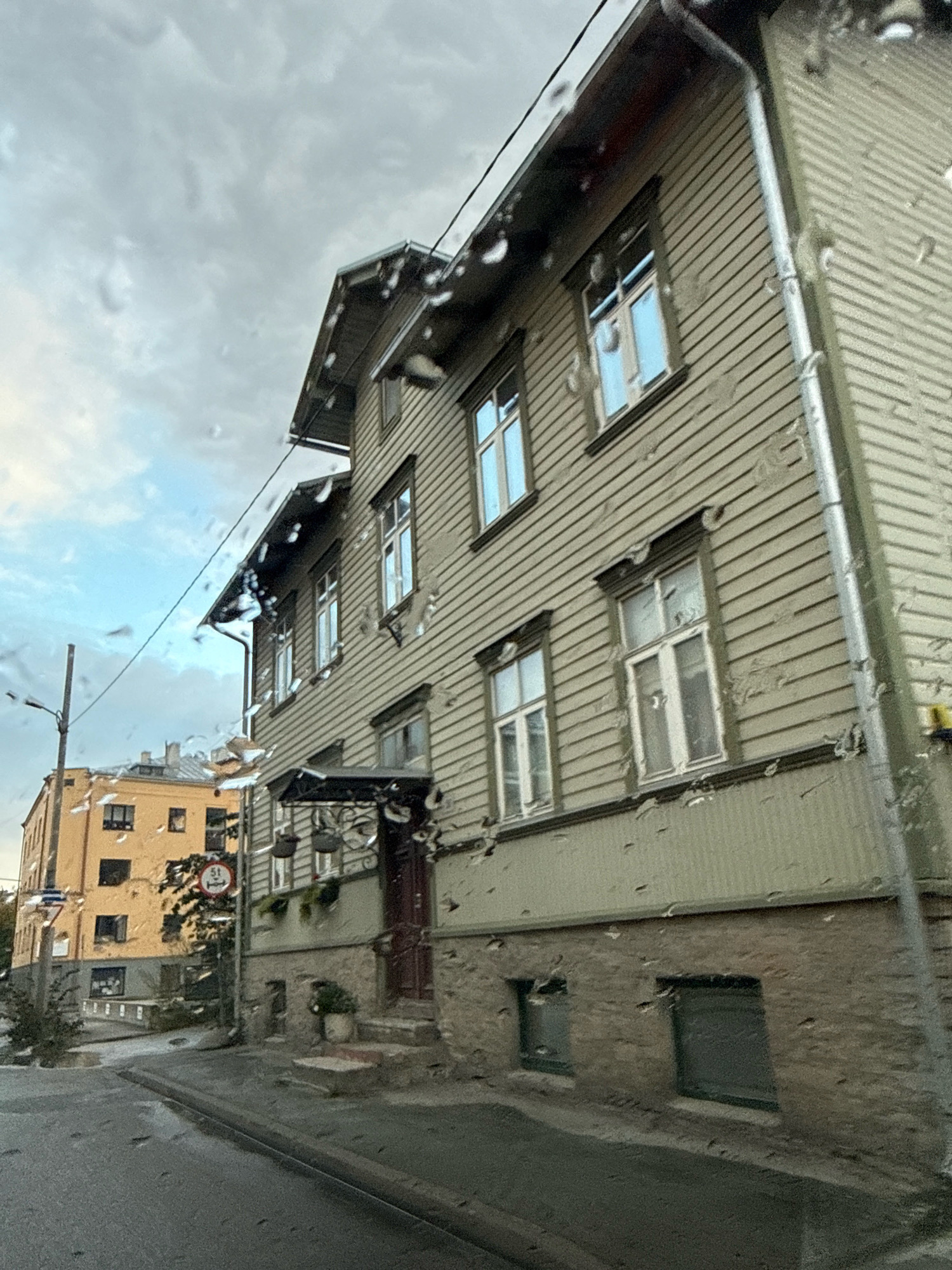 The image depicts a street scene in Tallinn, Estonia, on a cloudy, rainy day. In the foreground, raindrops are visible on the camera lens, obscuring some details. A two-story building with light green clapboard siding and a gray metal roof dominates the right side of the frame. It has multiple windows with white frames, some reflecting the sky. The building's foundation is made of stone. To the left of this building, a yellow apartment building with dark windows is visible. A street lamp with a visible pole stands on the sidewalk between the two buildings. A red and white circular traffic sign with a number "5t" is mounted on a pole. The street in front of the buildings is wet, indicating recent rainfall, and appears to be paved with asphalt. The sky is overcast with scattered clouds.