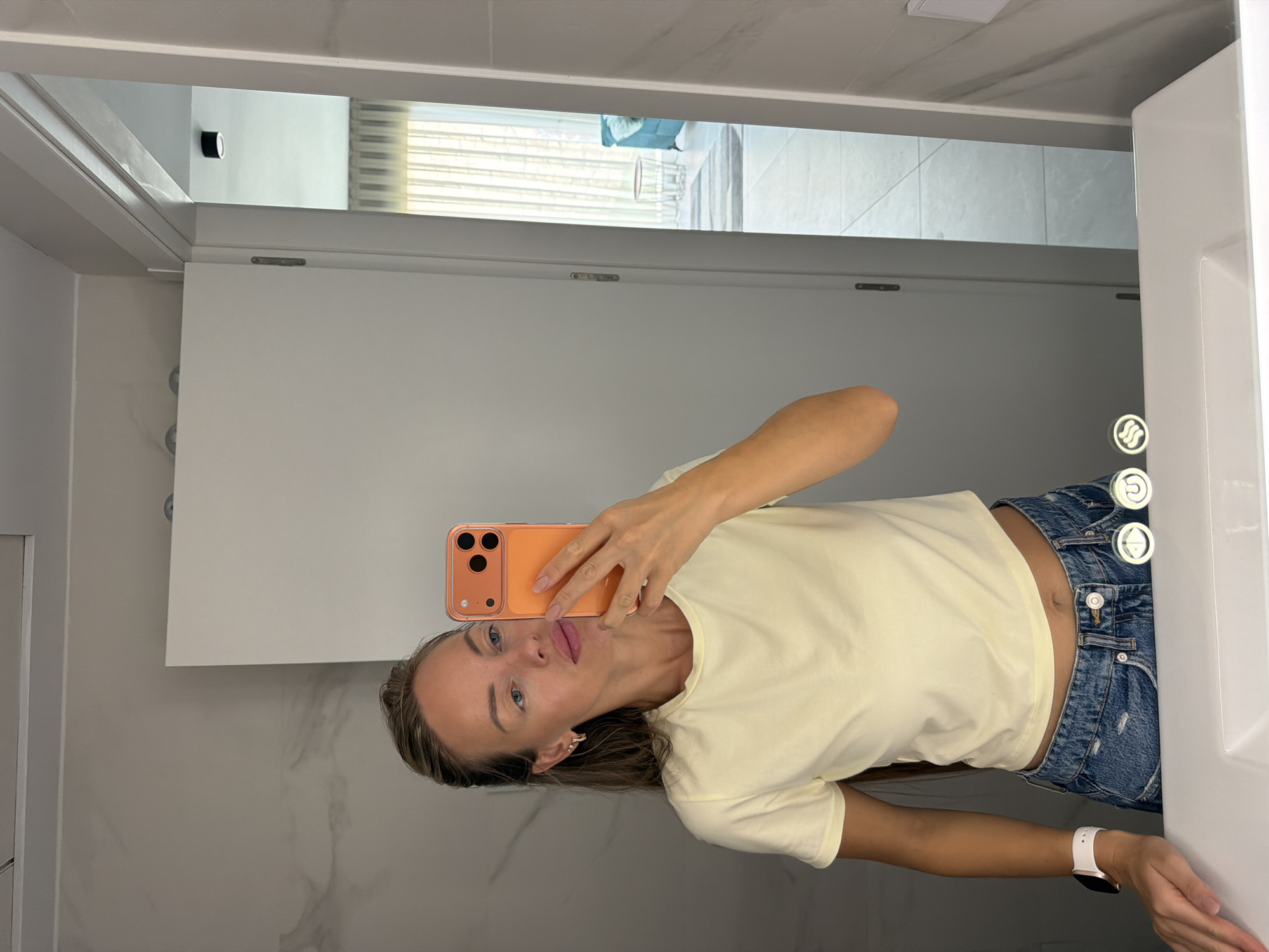 A woman takes a mirror selfie in a modern bathroom with marble-effect tiled walls. She is wearing a light yellow crop top and distressed denim shorts. Her right hand holds an orange smartphone, and her left hand rests on the edge of a white sink. A white smartwatch is visible on her left wrist. The background shows an open doorway leading to a brightly lit room with sheer curtains and a teal sofa.