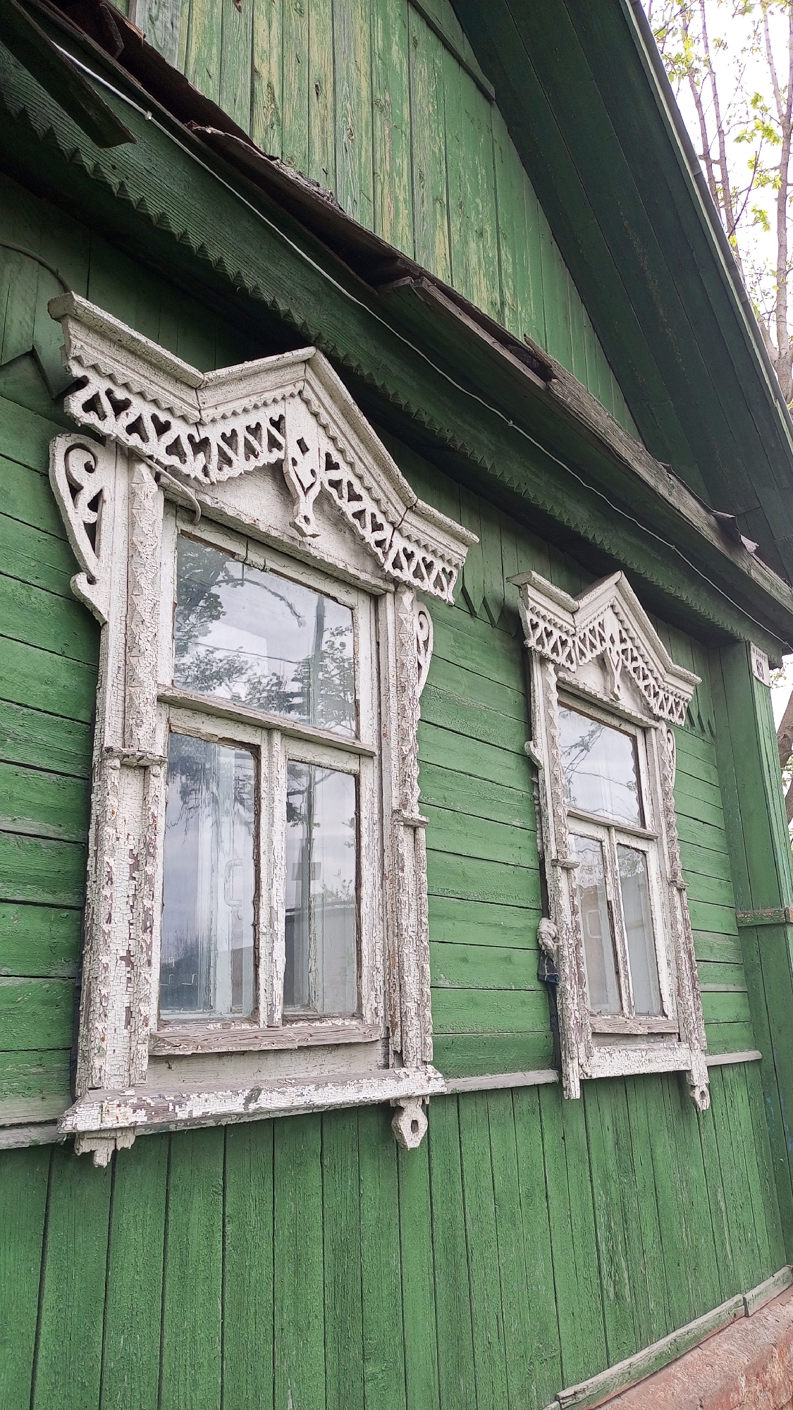 The image shows a section of a green wooden house in Юдино, Russia, featuring two windows with ornate, white, peeling trim. The house is constructed of vertical wooden planks, painted a faded green. The window frames are heavily decorated with carved details and covered in peeling white paint. The windows themselves are multi-paned. The lighting suggests an overcast day.