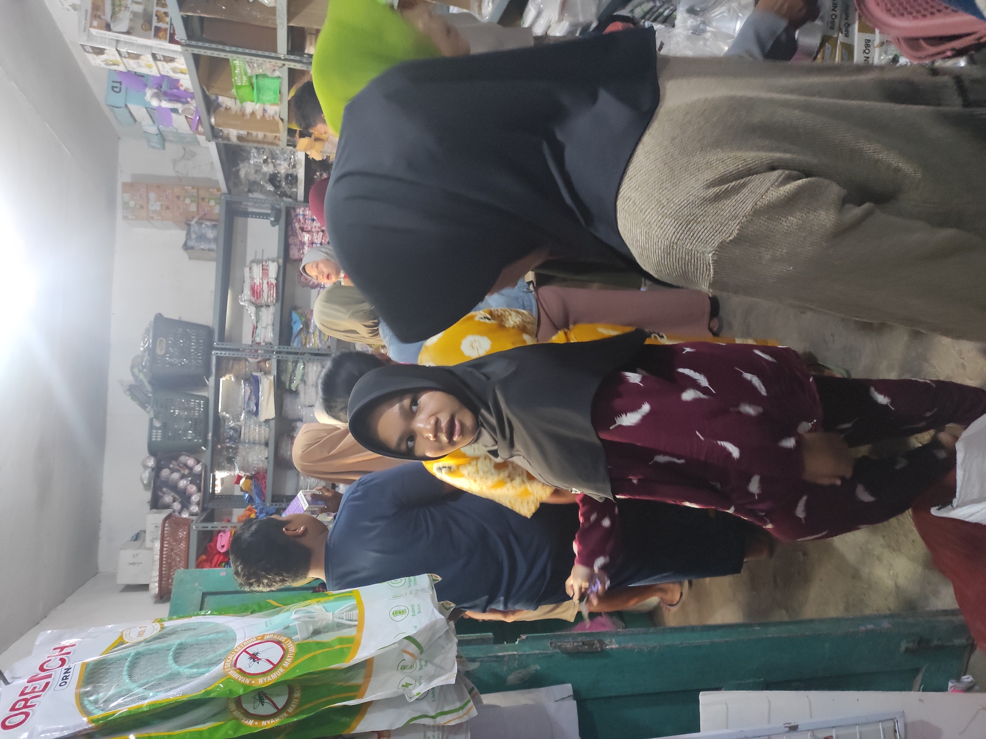 The image depicts an indoor scene at a small retail store or warehouse in Tempurejo, Indonesia, likely a general store or a supplier of household goods. The lighting is artificial and somewhat dim, characteristic of such establishments. In the foreground, to the left, a green vertical structure, possibly a door frame or a shelf unit, is visible. Hanging on the left side are several packages of mosquito swatters, branded "OREICH ORM," with green and white packaging featuring illustrations of mosquitoes and warnings.
The central area of the image shows a group of people, predominantly women, wearing hijabs, suggesting a cultural context common in Indonesia. One young woman in the middle ground, wearing a black hijab and a maroon outfit with a feather pattern, is looking towards the camera with a slightly surprised expression, holding a small object, possibly a receipt or a token, in her left hand. To her right, another person, only their back visible, is wearing a dark hijab and a beige knitted sweater. Other individuals are partially visible in the background, including a man in a dark blue shirt.
The environment is cluttered with shelves packed with various items. Shelves are filled with boxes, plastic containers, and other merchandise, indicating a retail or storage setting. Some shelves are made of grey metal, typical for such storage areas. In the background, more shelves are visible, stocked with a diverse range of products, including what appear to be stacked boxes of household goods, baskets, and possibly textiles. The floor is concrete and appears worn. The overall impression is of a busy, functional retail space.