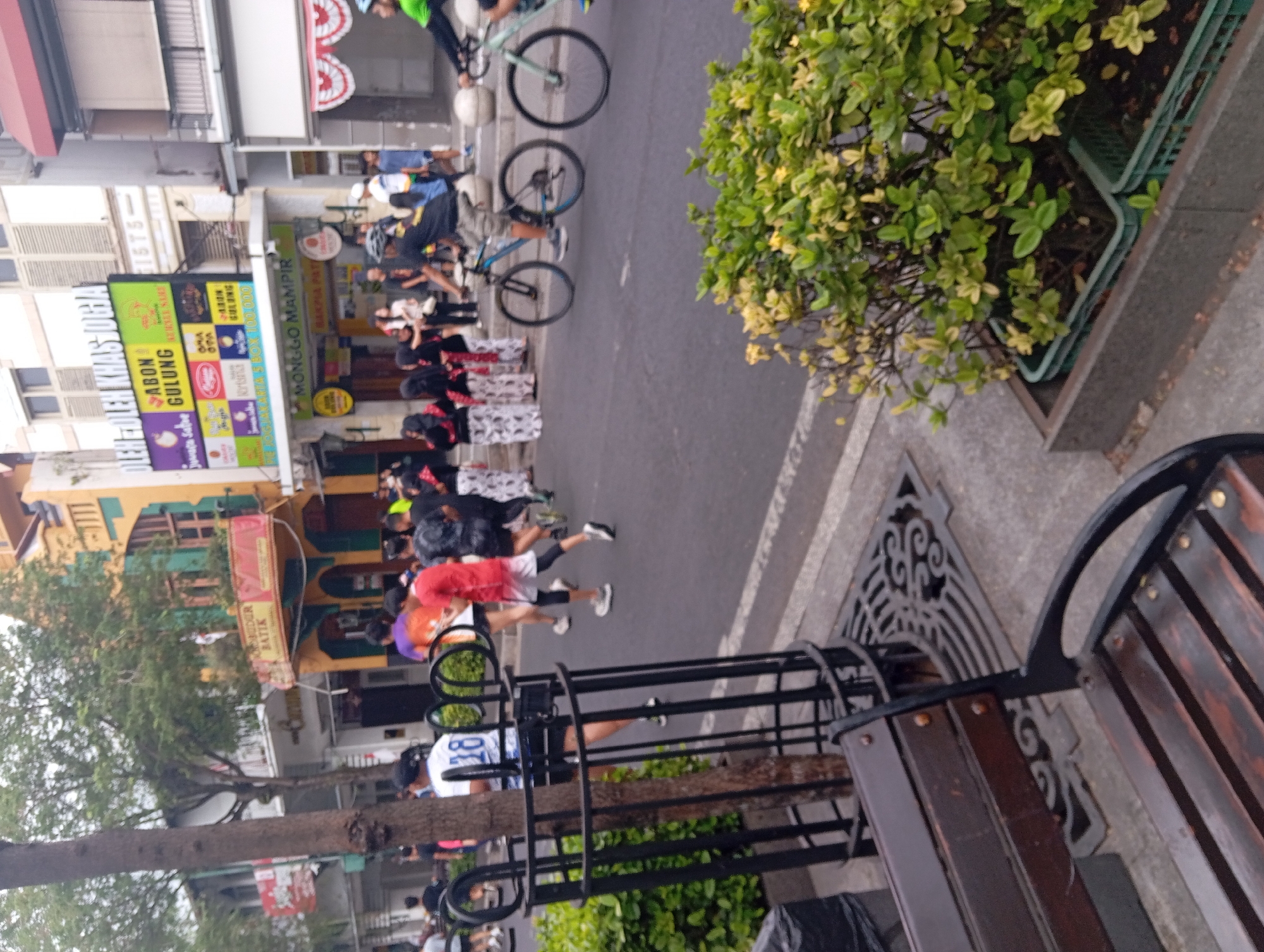 The image captures a street scene in the city of Yogyakarta, Indonesia, likely during a daytime event or market day. The foreground features a wooden bench and a decorative metal grate on the sidewalk. To the left, a mature tree stands next to a black metal enclosure.
The mid-ground shows a street with people participating in or observing an activity. Several individuals are running, including one wearing a white shirt with the number "18" on the back, and another in a red shirt. Others appear to be walking or standing. A group of women dressed in traditional batik attire, with black tops and patterned white skirts, are standing together. Two men are on bicycles, one on a blue bicycle and the other on a light green bicycle.
The background reveals the urban environment of Yogyakarta. Buildings line the street, with visible signage for shops. One prominent sign reads "OLEH OLEH KHAS JOGJA" (Souvenirs from Yogyakarta) and includes various product advertisements. Another sign says "MONGGO MAMPIR" (Please Come In). The architecture suggests a mix of traditional and modern styles, with colorful facades and shutters. Green foliage is visible in planters along the street and on balconies. The overall atmosphere appears to be lively and bustling.