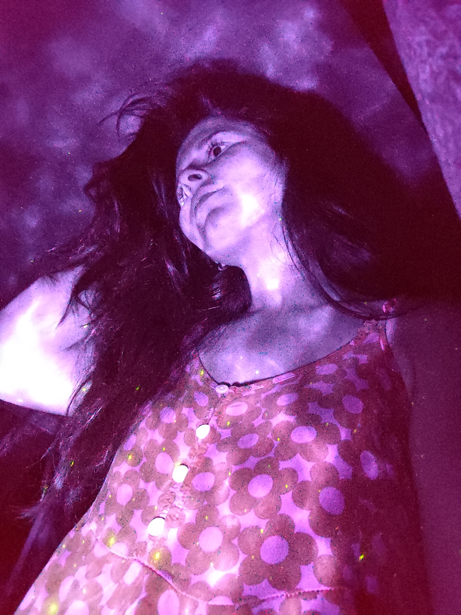 A woman with long dark hair is looking upward. She is wearing a pink dress with a floral pattern and buttons down the front. The lighting is purple and red. The photo appears to be taken from a low angle. The location is unknown and cannot be identified as Фоки, Russia.