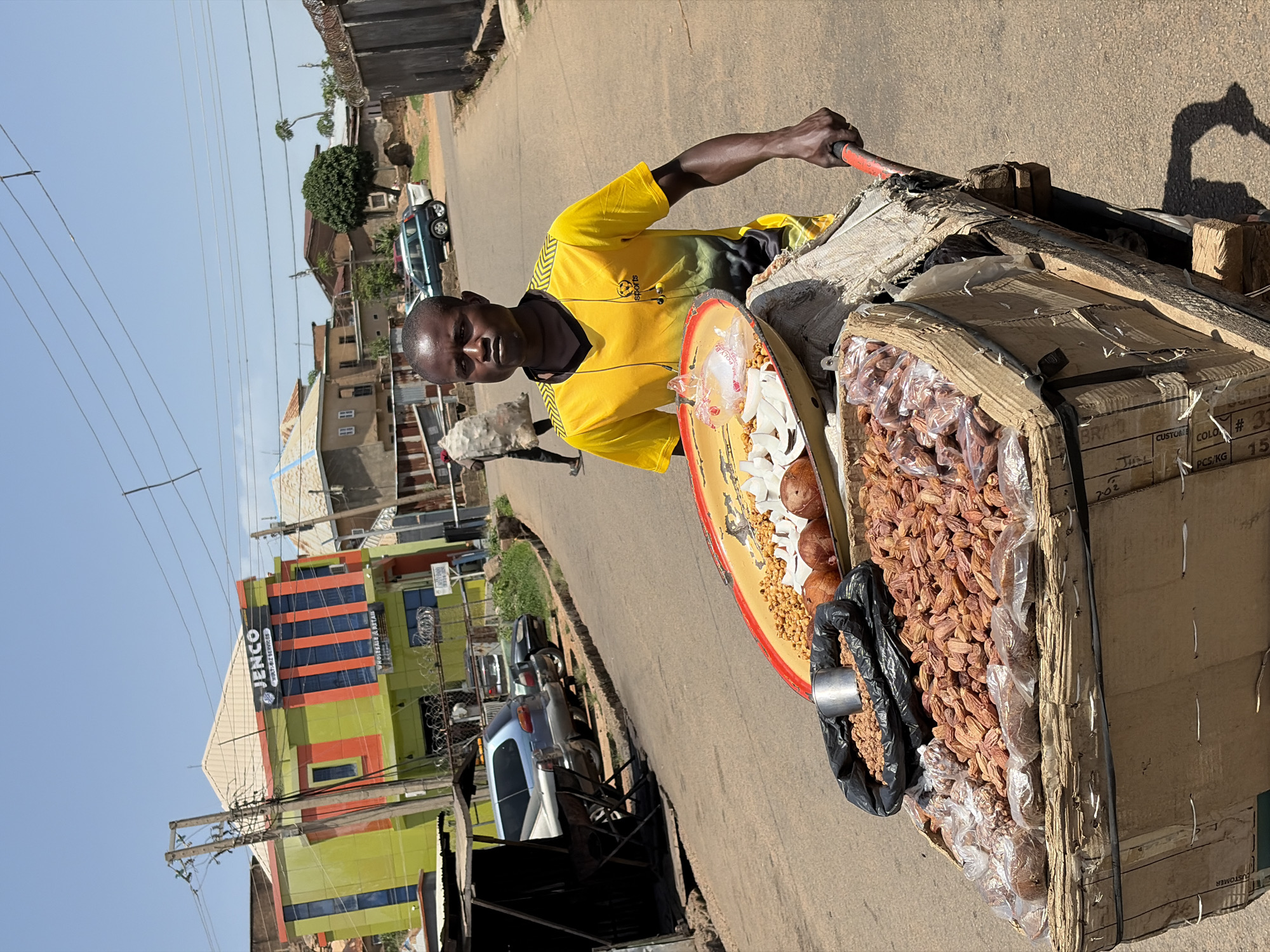 A vertical photograph, rotated 90 degrees counter-clockwise, captures a street vendor with his mobile cart on a sunny day in Jos, Nigeria.
The main subject is a man of dark complexion, wearing a yellow and black short-sleeved t-shirt with "sports" visible on the chest. He stands beside his cart, looking directly at the camera with a neutral expression, holding onto a red handle. His cart is laden with goods for sale. On a large, round, red-rimmed tray, there are white chunks of coconut, small light brown nuts (likely peanuts or groundnuts), and several whole brown coconuts. Next to the tray, a cardboard box is filled with numerous individually packaged dried fruits, possibly dates. A black plastic bag containing more goods, with a metal measuring cup resting inside it, is placed within the box. Text visible on the cardboard box includes "CUSTOMER", "702", "BRATO", "CUSTOME COLO #33", "PCS/KG", and "15".
The setting is an asphalt road, likely a street or market area. In the background, there are various buildings under a clear, bright blue sky. A prominent two-story building features green and orange sections with vertical blue stripes, and a sign that reads "JENCO MULTISERVICES" with partially visible text "WHOLESALE & RETAR" below it. Power lines crisscross the upper portion of the frame. Several vehicles are parked along the street further down. Another person carrying a large white sack is visible walking in the mid-ground. The strong sunlight casts clear shadows, indicating it is likely midday.