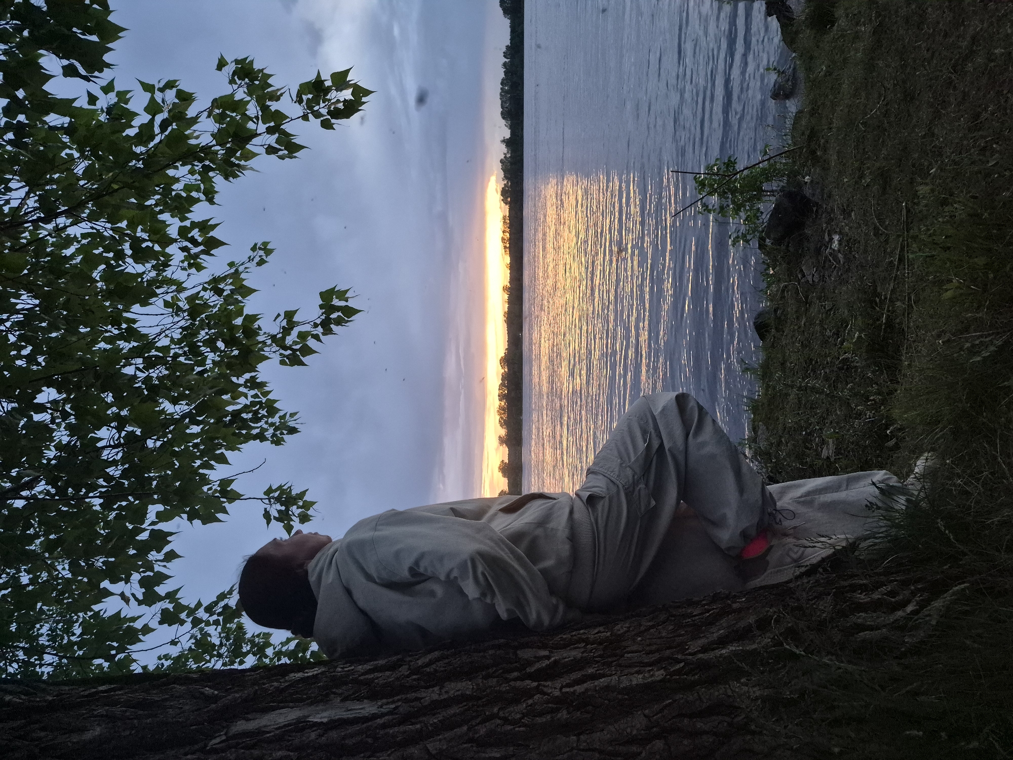 A person lies on a tree trunk near a body of water at twilight, presumably in the city of Chaikovsky, Russia. The water reflects the sunset, and the person is dressed in light-colored clothing. Branches of a tree with dense foliage are visible to the left. Grass and vegetation are visible on the right side of the image.