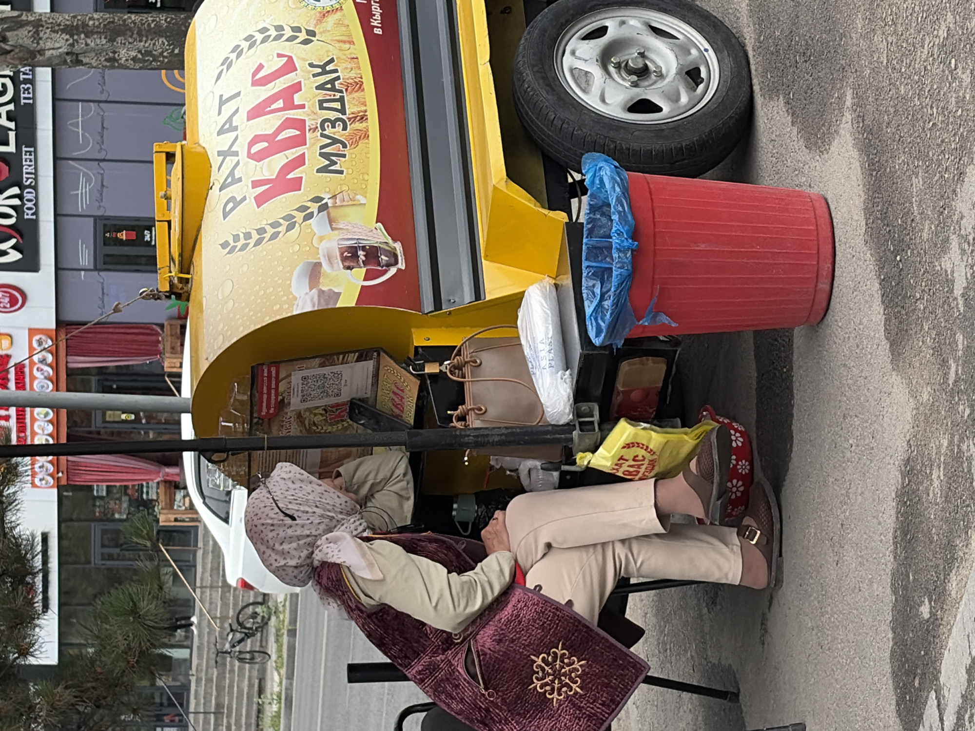 The image captures an outdoor scene in an urban environment, likely a street or sidewalk. In the foreground, a woman is seated on a dark chair, facing to the left. She is wearing a light-colored headscarf with a patterned print, a light-colored long-sleeved shirt, and a dark maroon vest adorned with intricate gold embroidery. Her lower half is covered by light-colored pants, and she is wearing brown slip-on shoes. Her right arm is resting on a surface, and her head is tilted downwards.
Beside her, to her right, is a bright yellow mobile kiosk or cart. The side of the kiosk features prominent text and imagery related to "KVAS" and "MUZDAK," with depictions of wheat stalks and frothy mugs of beer. A red trash can with a blue plastic liner is situated in front of the kiosk, to the right of the woman. Various items, including a yellow plastic bag and a light-colored handbag, are placed around the kiosk and near the woman.
In the background, a white car is parked on the street. Further back, a building with large glass windows and storefronts is visible, with red curtains partially obscuring the view. A bicycle is parked on a stepped area to the left of the car. Trees with green foliage are present, casting shadows on the ground. The overall setting appears to be a casual commercial or service area within a city.