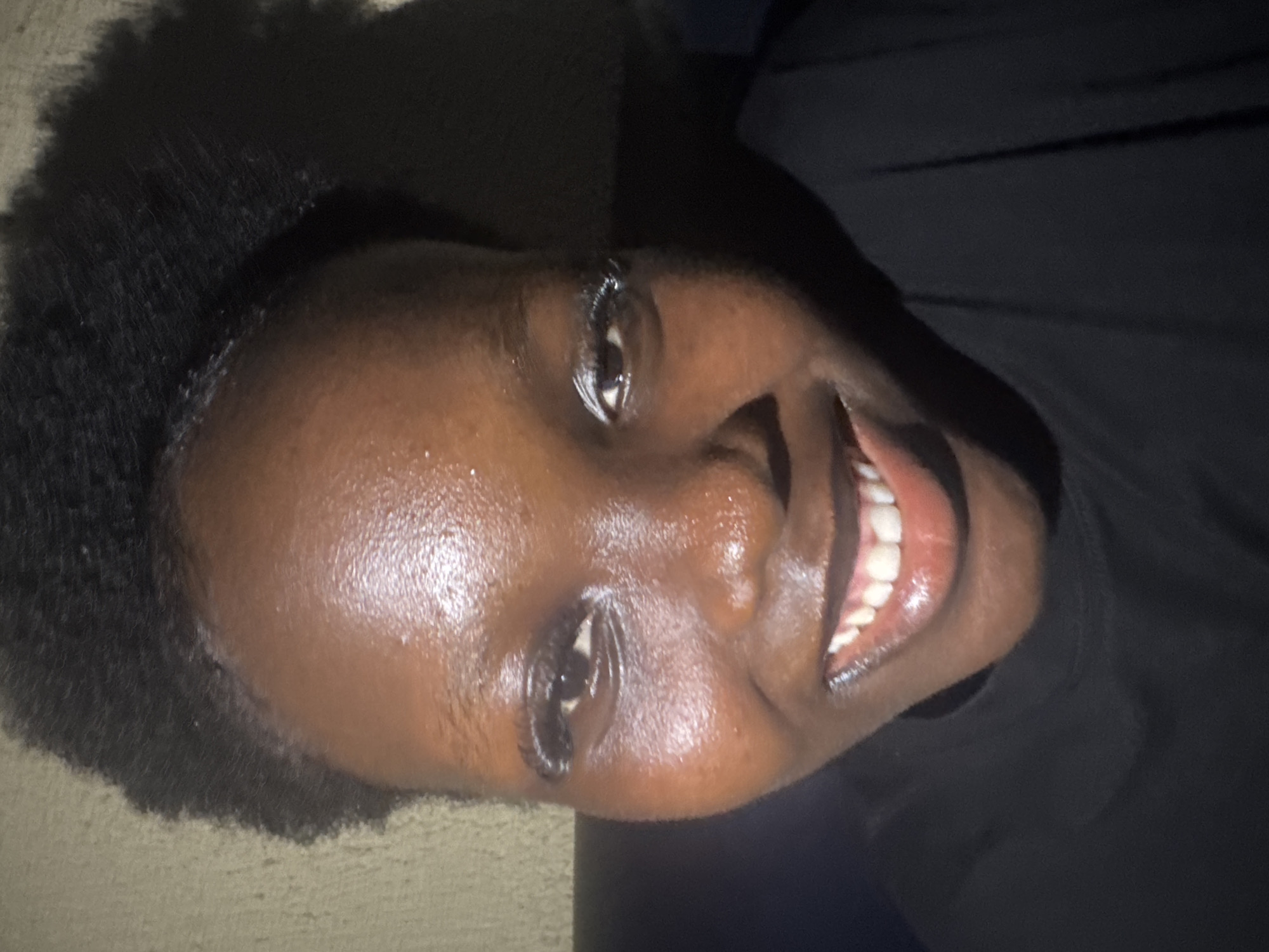 A close-up, rotated image shows a person with dark skin and short, dark hair. The person is smiling, revealing their teeth. They appear to be wearing a dark-colored garment. There is no contextual information to definitively determine the location as Onitsha, Nigeria, or any other specific place.