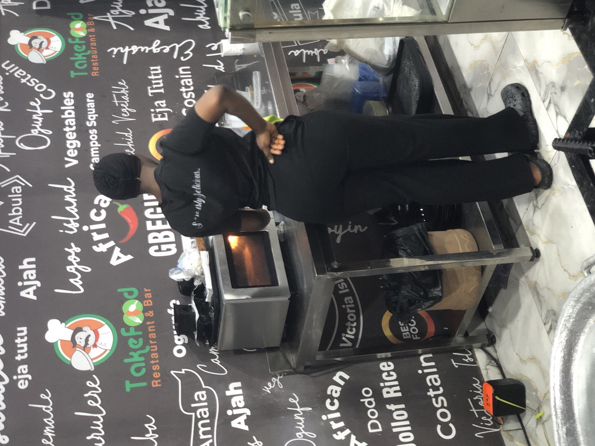 This indoor photograph captures a staff member at what appears to be a Nigerian restaurant or food establishment. The primary subject is a person, seen from behind, wearing a black uniform including a black t-shirt, trousers, and a head covering, along with black Crocs. They are standing behind a stainless steel counter or food warmer, with their hands positioned on their lower back, suggesting a moment of pause or slight strain. Inside the warmer, a warm orange glow indicates it is active.
The setting is characterized by a dark wall serving as a menu board or branding display, covered with white handwritten-style text and a prominent logo. The logo features a chef with a mustache and hat, holding a covered dish, alongside the name "TakeFood Restaurant & Bar." Below the counter, various items are stored, including black plastic bags and brown paper bags, possibly for packaging orders. The floor is tiled with a light-colored, marbled pattern.
Visible text on the wall includes "TakeFood Restaurant & Bar" (appearing multiple times), and several Nigerian food items and locations such as "Amala," "Ajah," "Eja Tutu," "Abula," "Ogunfe," "Costain," "Lagos island," "Vegetables," "African," "Campos Square," "Elegushi," "Dodo," "Jollof Rice," and "Victoria Island." The person's shirt also has partially visible text "S...tly delicious." Another logo seen on items below the counter reads "BES FOC." The scene depicts a typical busy kitchen or serving area in Ado, Nigeria.