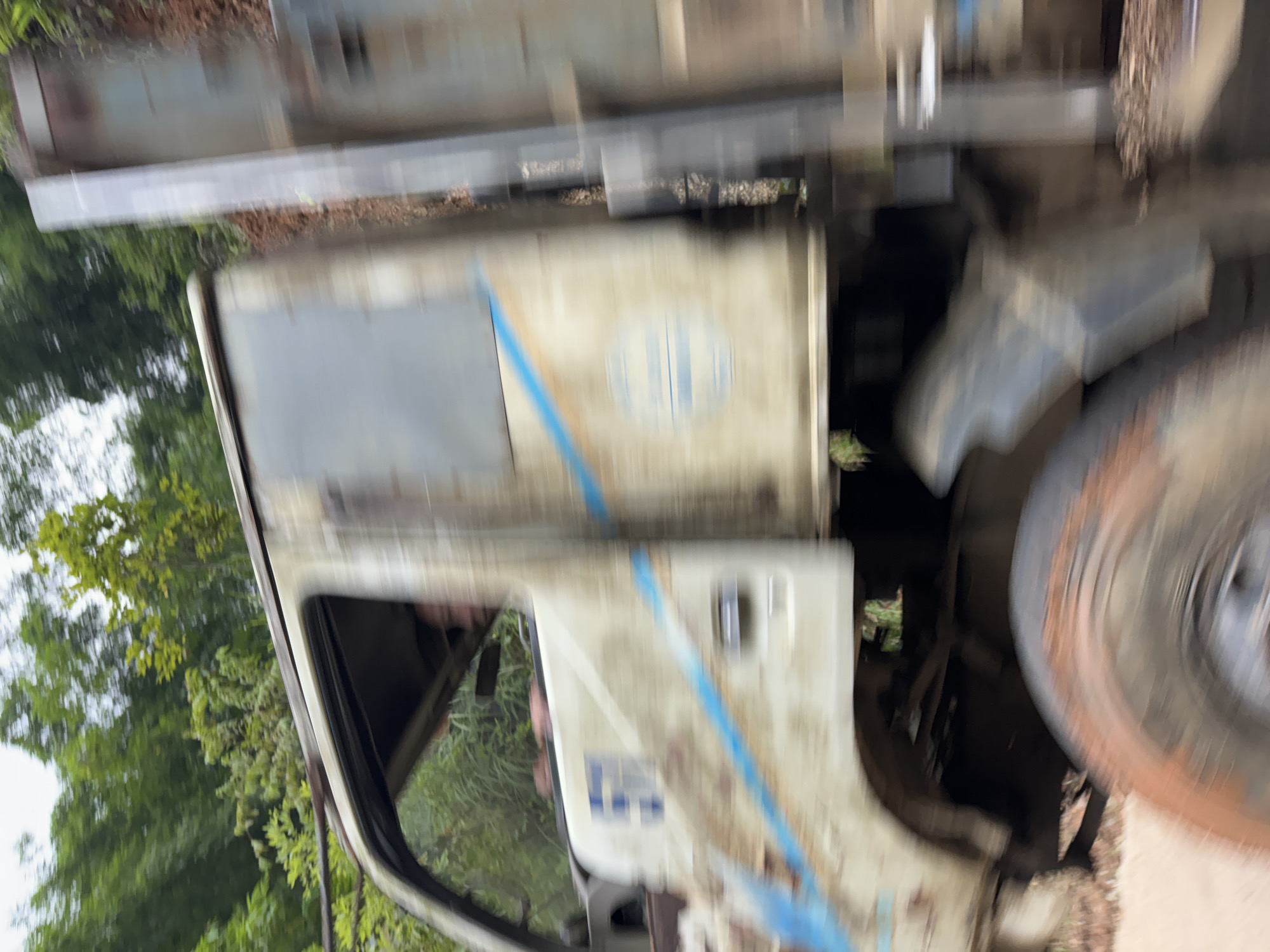 The image captures a blurry, motion-filled scene of a weathered, light-colored truck driving on a dirt road. The truck is old and appears to be in disrepair, with rust and dirt visible on its body. A blue stripe runs diagonally across the side of the truck's cab. A person is partially visible inside the driver's side of the cab, with their hands on the steering wheel. The truck is surrounded by lush green foliage, with trees and vegetation visible on either side of the road. The ground is reddish-brown, typical of a dirt or clay road. The motion blur suggests the truck is moving at a significant speed. The overall impression is of a rural or remote setting, likely in Cổ Lũng Commune, Vietnam, given the vegetation and road conditions.