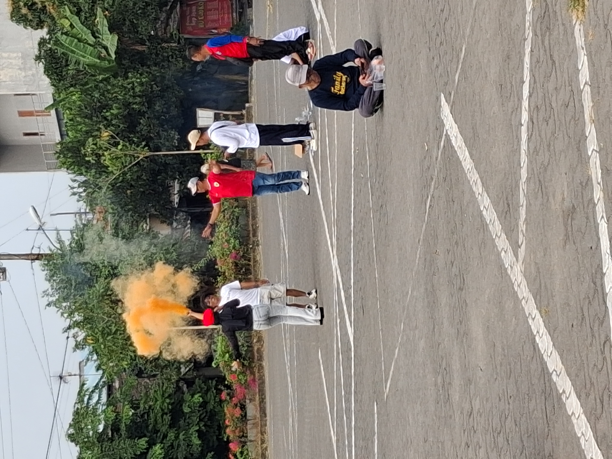 In Surakarta, Indonesia, a group of people gather in an outdoor paved area, likely a parking lot or sports court, marked with white lines. A person in the foreground, seated on the ground, is looking at a phone. Further back, a person in a black hijab and red headscarf holds a smoking orange flare, facing away from the camera. Another person in a white shirt and shorts stands nearby, appearing to interact with the person holding the flare. A man in a red shirt and jeans stands to the left, with his arm extended. To the right, two men in athletic attire are also present, one in a white long-sleeved shirt and the other in a red and blue shirt. Trees and buildings are visible in the background, suggesting an urban environment. The sky is overcast.