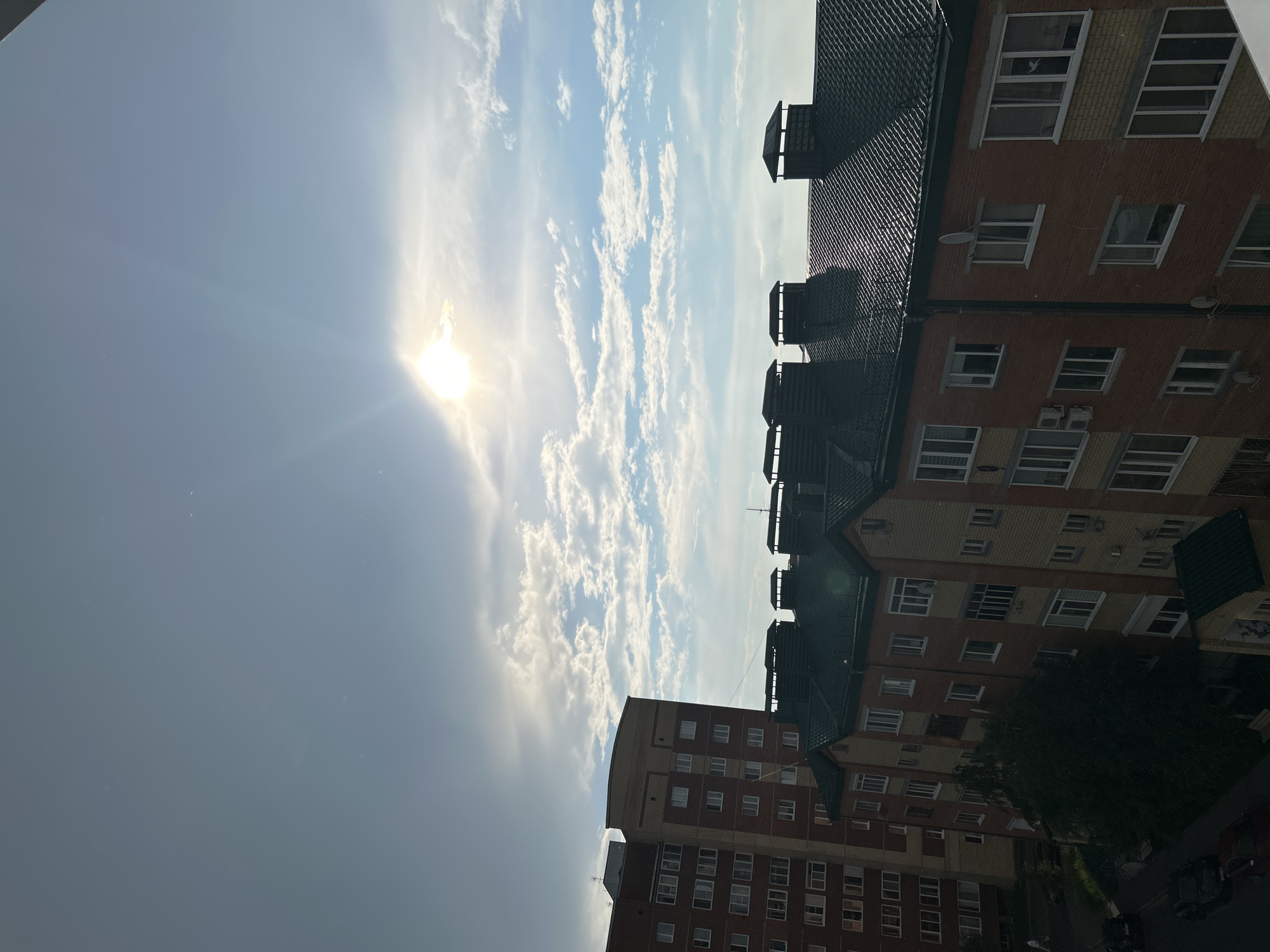 This media file is a vertical, outdoor, daytime photograph taken from a high vantage point, likely a window or balcony, looking up at the sky and the sides of apartment buildings. The sun is prominently visible, creating a lens flare and casting a bright light across the scene. Wispy white clouds are scattered across a blue sky, indicating fair weather.
Two apartment buildings are the main objects in the scene, their brick facades and numerous windows clearly visible. The buildings are modern, with dark, sloped roofs and what appear to be air conditioning units or vents on some of the walls. The architecture suggests a residential area. There are dark green roofs on the buildings closest to the viewer, and a large, dark green tree is visible between the buildings, suggesting a park or green space nearby.
The time of day is clearly daytime, with the sun high in the sky. The lighting suggests it could be morning or afternoon, given the angle of the sun and the bright glare. There are no people visible in the image. The setting is urban, specifically a residential area in Солнечный, Russia. There is no visible text in the image. The overall mood is bright and somewhat serene, dominated by the natural elements of the sun and sky.