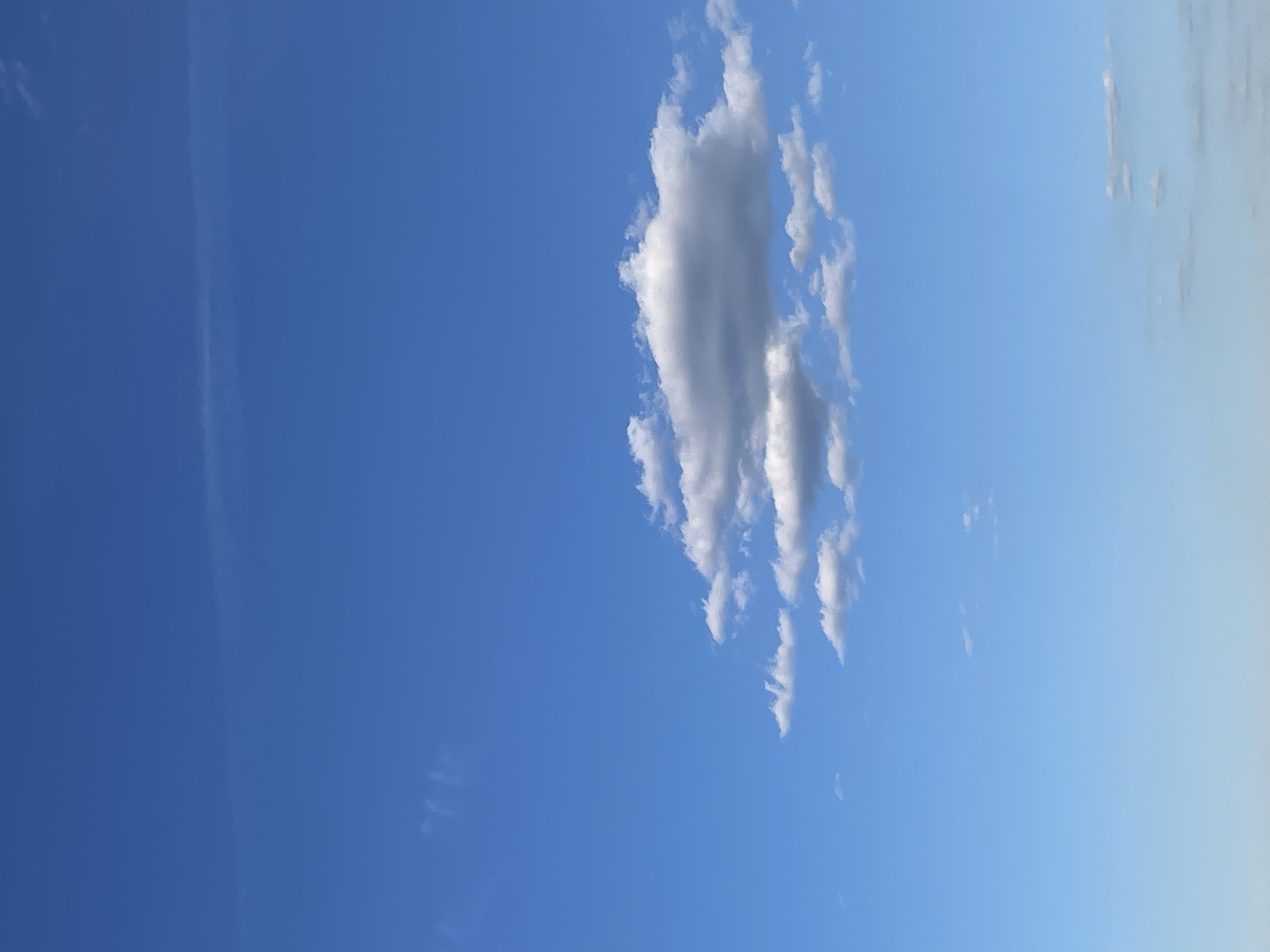 A low-angle shot captures a vibrant blue sky, with a single, vertical arrangement of puffy white clouds dominating the scene. To the left, a faint, linear contrail stretches across the sky. The blue gradient shifts slightly towards the right, indicating a subtle change in atmospheric density. The location is Unknown, Unknown.