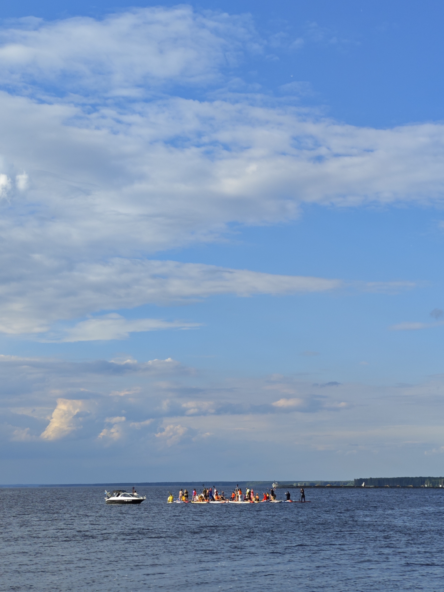 In this medium shot, a large group of people are enjoying a summer day on the water. The scene is set on a calm, dark blue body of water under a bright blue sky dotted with fluffy white clouds. In the foreground, a small, white motorboat with a black canopy is moving across the water. Behind the motorboat, a much larger group of people are gathered on several paddleboards or a makeshift raft, some of whom are standing and appear to be posing for the photo. A few individuals are in the water, seemingly playing or swimming. The activity appears to be a recreational outing, possibly a group paddleboard event or a gathering of friends. The overall atmosphere seems relaxed and enjoyable. In the background, a long, dark line of trees marks the distant shore, with a faint hint of a structure or pier. The lighting suggests it is daytime, likely late afternoon given the quality of the light and the sky. There is no visible text in the image.