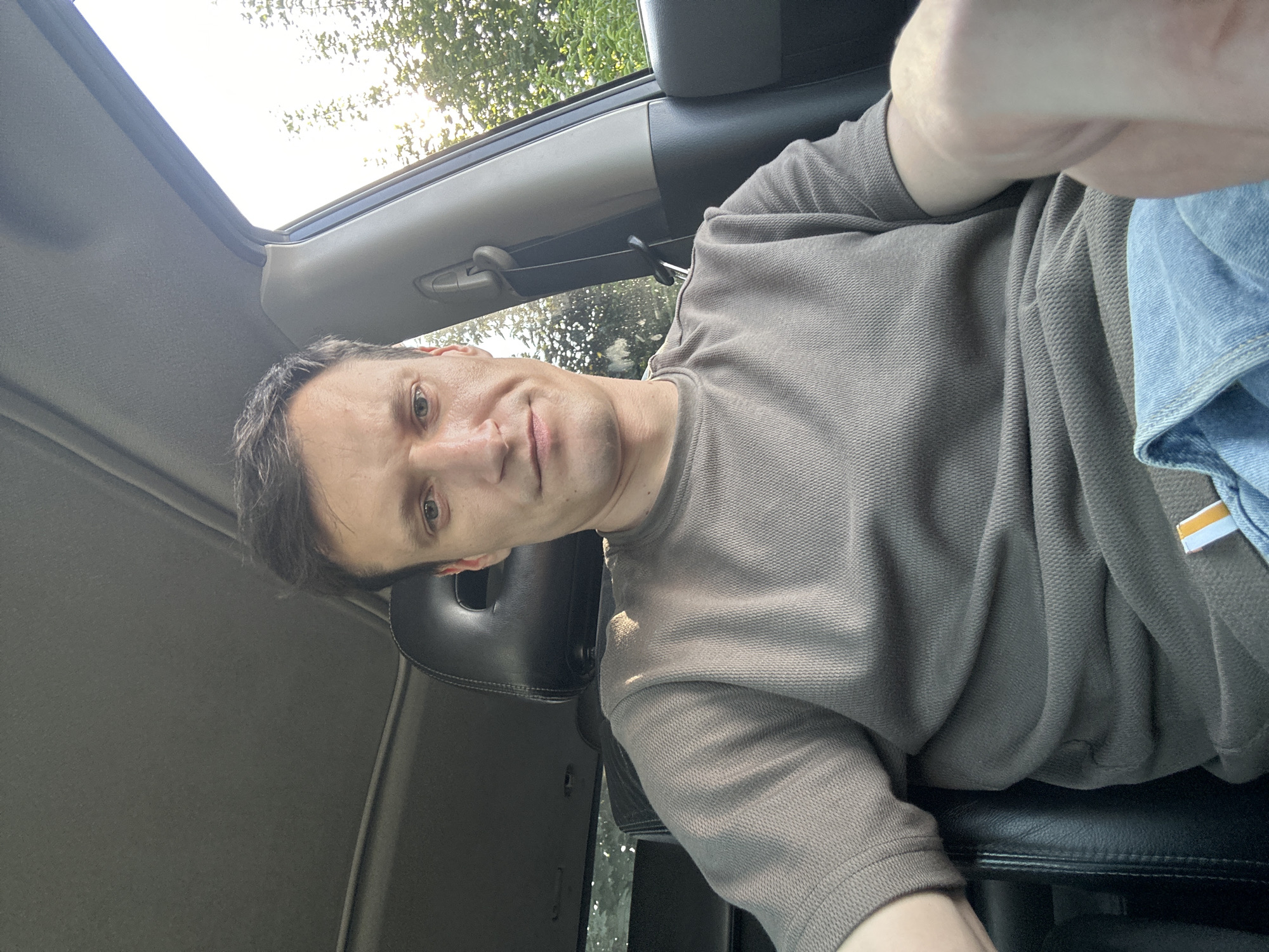 A man is sitting in the driver's seat of a car, facing forward and looking at the camera. He has short, dark hair and light skin. He is wearing a grey t-shirt and blue jeans. The interior of the car is visible, including the dashboard, headrest, and seatbelt. Sunlight is coming in through the window, illuminating the scene and casting shadows. The car appears to be parked outdoors, with trees and foliage visible outside the window. The location is the city of Shatava, Ukraine.