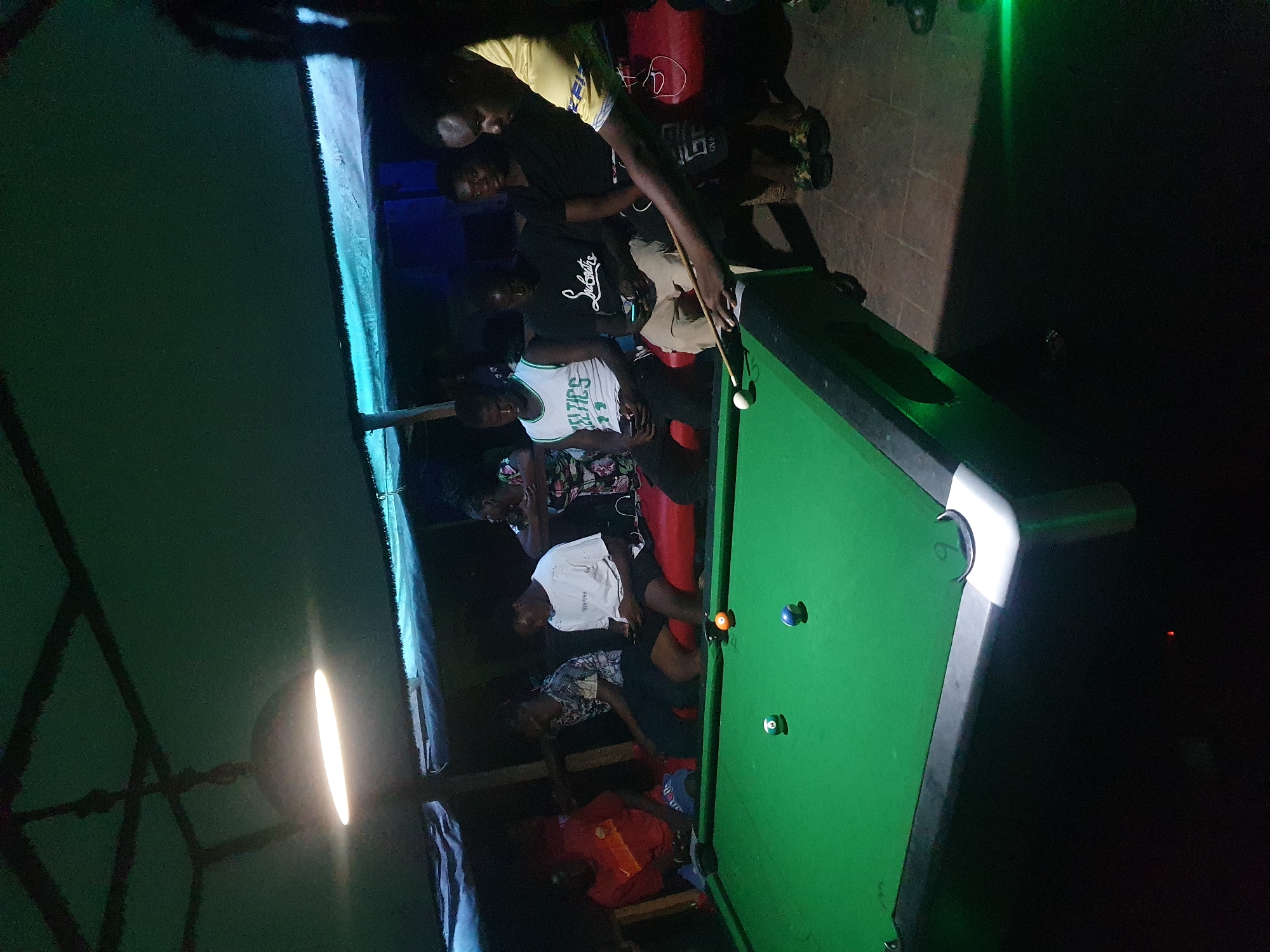 The image depicts a lively night scene at a recreational establishment in Lokoja, Nigeria, likely a bar or game room. A group of young men, approximately eight to ten individuals, are gathered around a green-felt pool table. One person is actively playing, holding a pool cue stick and lining up a shot at the white cue ball. An orange 5-ball, a blue 2-ball, and a green 6-ball are visible on the table.
Other individuals are standing or sitting around the table, watching the game or socializing. Notable attire includes a white jersey with "CELTICS" printed on it and a black t-shirt with "Louboutin" partially visible. The atmosphere appears relaxed and social.
The setting is illuminated by several artificial light sources, indicating it is nighttime. A bright, elongated light fixture hangs from the ceiling, directly above the table. A distinct blue glowing light strip runs along a vertical structure in the background, adding to the ambient lighting, and some green light can be seen further back. The floor consists of paving stones or tiles. The scene conveys a sense of casual enjoyment and camaraderie among the participants.