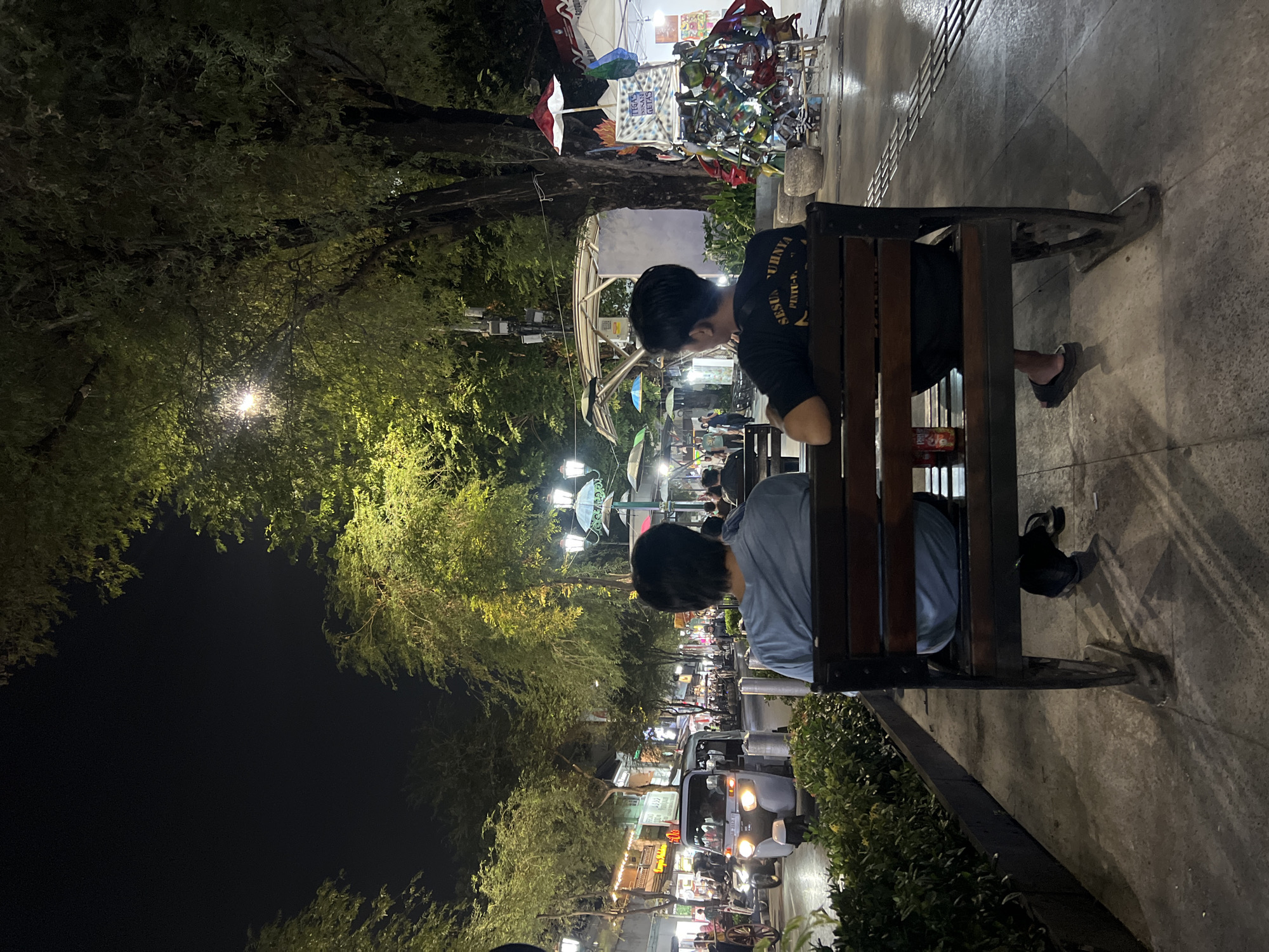 The image captures a nighttime scene on Malioboro Street in Yogyakarta, Indonesia. Two young men are seated on a wooden bench, facing away from the camera. The man on the left wears a light blue t-shirt, and the man on the right wears a black t-shirt with text on the back. A red beverage can sits on the bench between them. The street is illuminated by overhead lamps and the lights of shops and passing vehicles. A small motorized vehicle, resembling a tuk-tuk, is visible in the street along with other traffic. Trees line the sidewalk, their leaves casting shadows. In the background, market stalls and shops are open, and people are walking around. The overall atmosphere is that of a bustling urban environment at night.