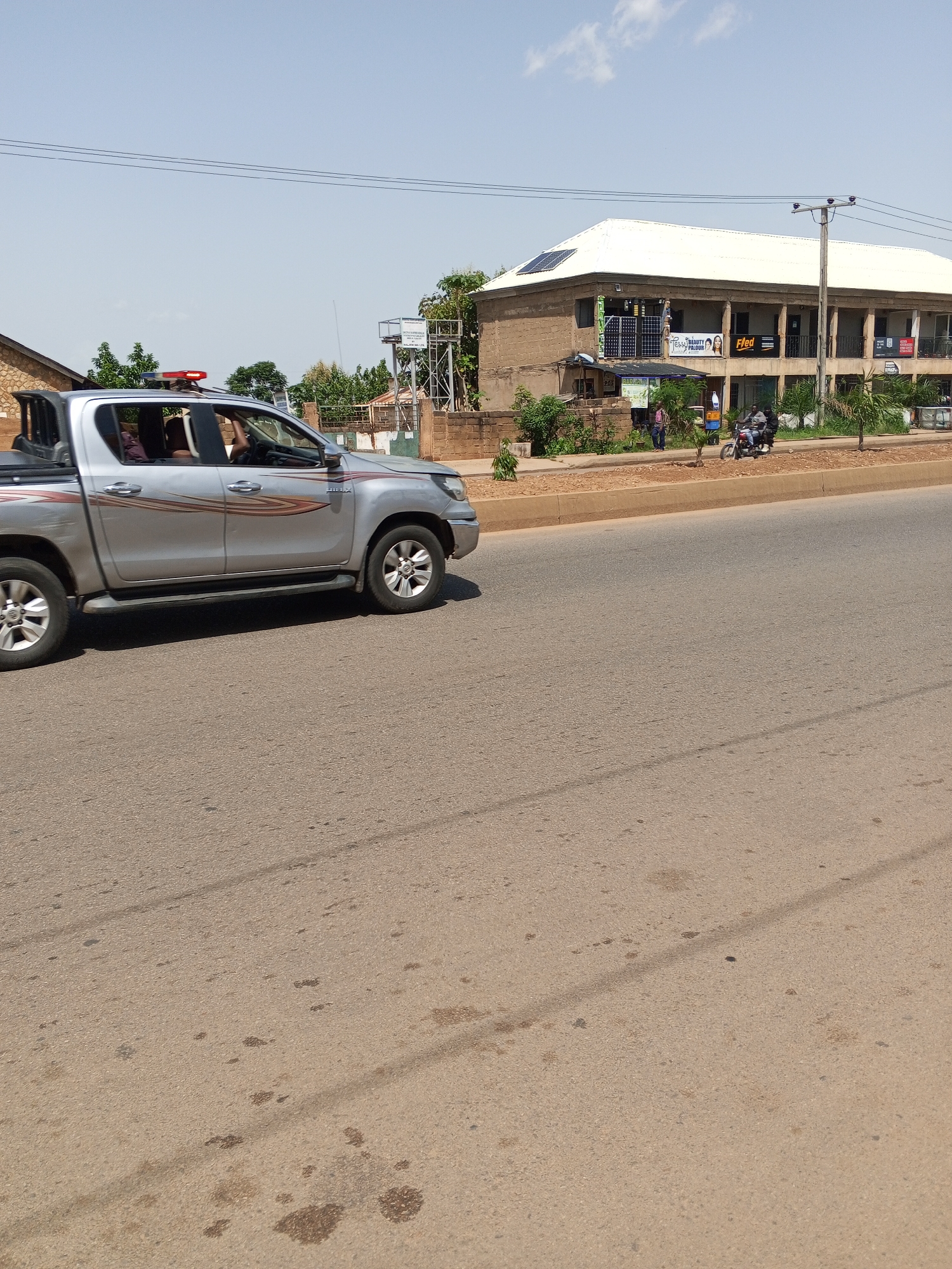 The image shows a silver Toyota Hilux pickup truck with a red light bar on its roof, moving on a paved road. The truck has a decal with red and orange stripes on its side. In the background, there is a two-story building with a white roof, solar panels, and several businesses on the ground floor. Two people are visible on a motorcycle in front of the building, and another person is standing on the sidewalk. The environment appears to be an urban or semi-urban setting in Bukuru, Nigeria, with a clear sky and some greenery. The road surface is asphalt and shows signs of wear and some oil stains.