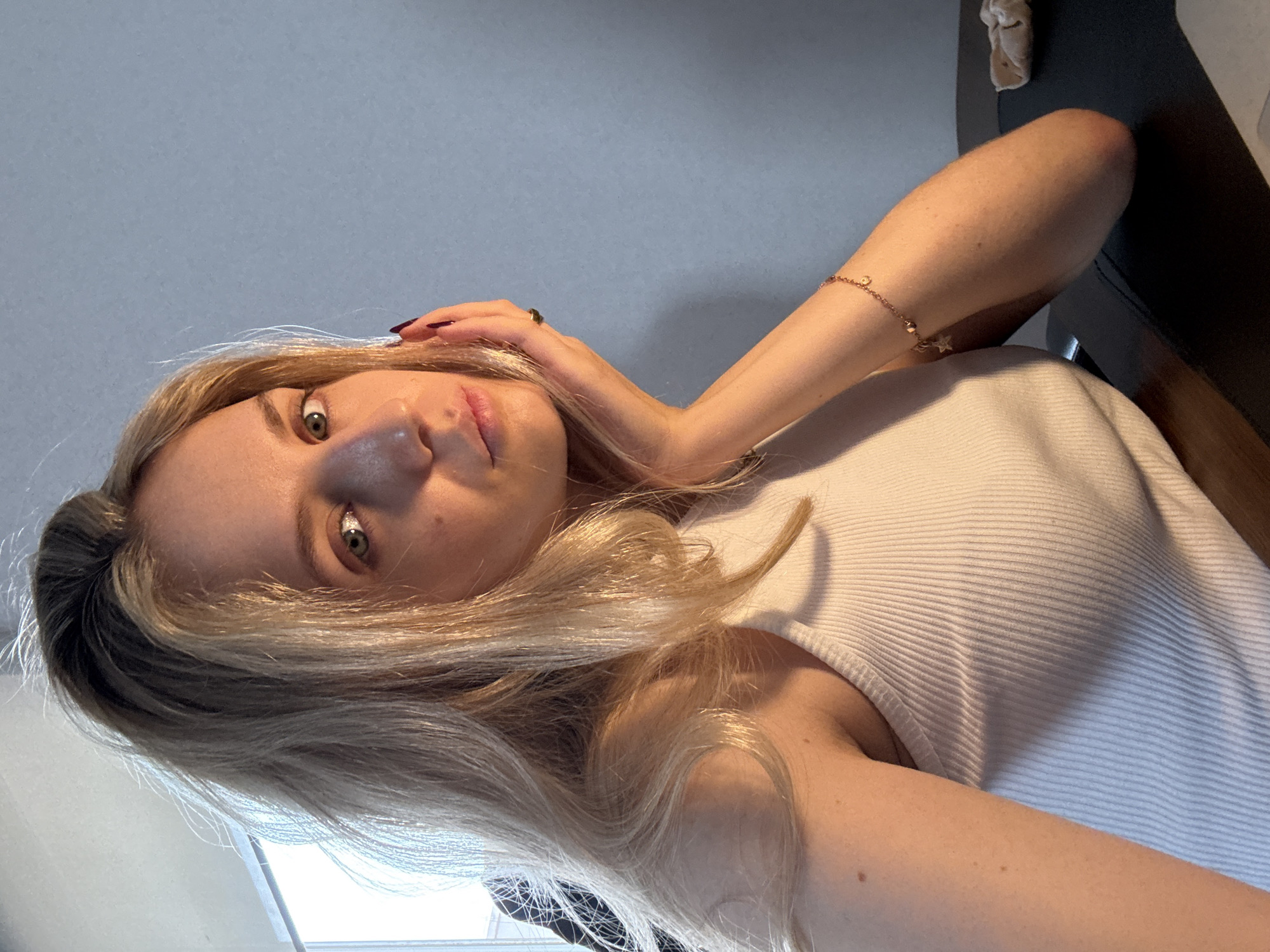 A fair-skinned young woman with light blonde hair and green eyes is facing the camera. She is wearing a white ribbed tank top. A gold bracelet adorns her left wrist. Her left hand is near her face, and she has dark nail polish on her fingernails and a ring on her finger. The background appears to be a neutral grey wall. A dark object is visible in the upper right corner. There is no evidence to determine if the location of the scene is in Mexico City, Mexico.