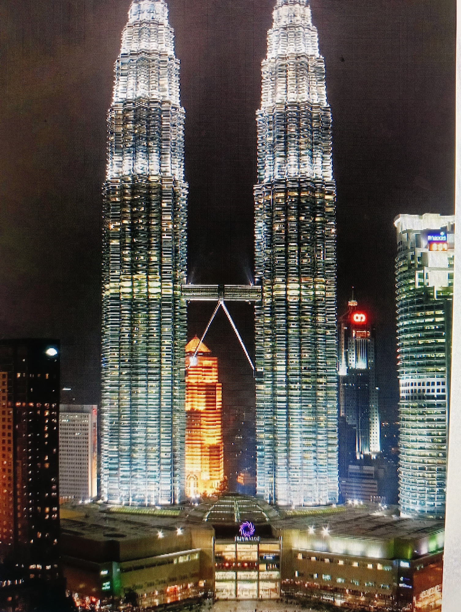 The image depicts a night view of the Petronas Twin Towers in Kuala Lumpur, Malaysia. The iconic skyscrapers are illuminated with artificial lights, highlighting their intricate architectural details. A skybridge connects the two towers at a mid-level. In the foreground, there are other buildings and structures, also lit up, suggesting a bustling urban environment. The sky is dark, indicating it is nighttime. The image does not show the city of Sluck, Belarus; it is a depiction of Kuala Lumpur.