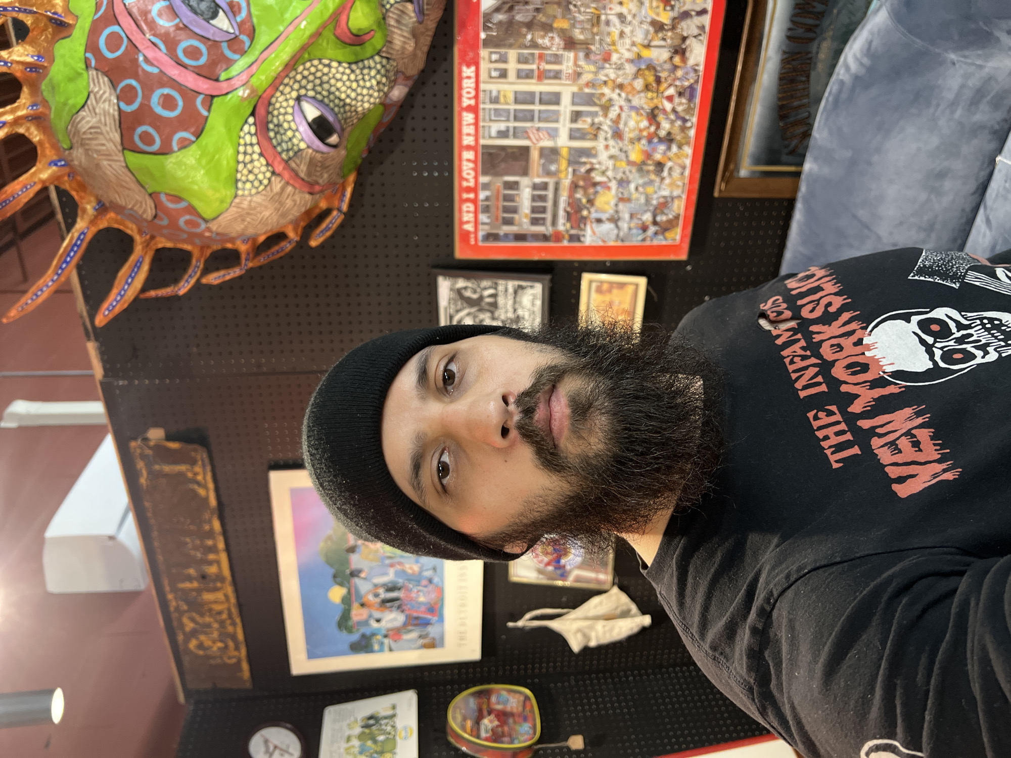 The image shows a person with dark hair and a beard wearing a black beanie and a black long-sleeved shirt with a skull design and red lettering that reads "THE INFAMOUS NEW YORK SLICE". The person is positioned in front of a wall adorned with various objects.
On the wall, there is a large, colorful papier-mâché mask or sculpture in the upper left corner. To the right of it, a framed poster or print titled "...AND I LOVE NEW YORK" is visible. Other framed pictures and decorative items are also hanging on the wall, including a rectangular wooden sign, a poster with an illustration, a white face mask, and a small tin can. The wall appears to be a display or shop area.
The location is suggested by the "I LOVE NEW YORK" print and the "NEW YORK SLICE" text on the shirt, but the specific city of City of Newburgh, United States of America is not directly evident from the visual content of this image alone.
