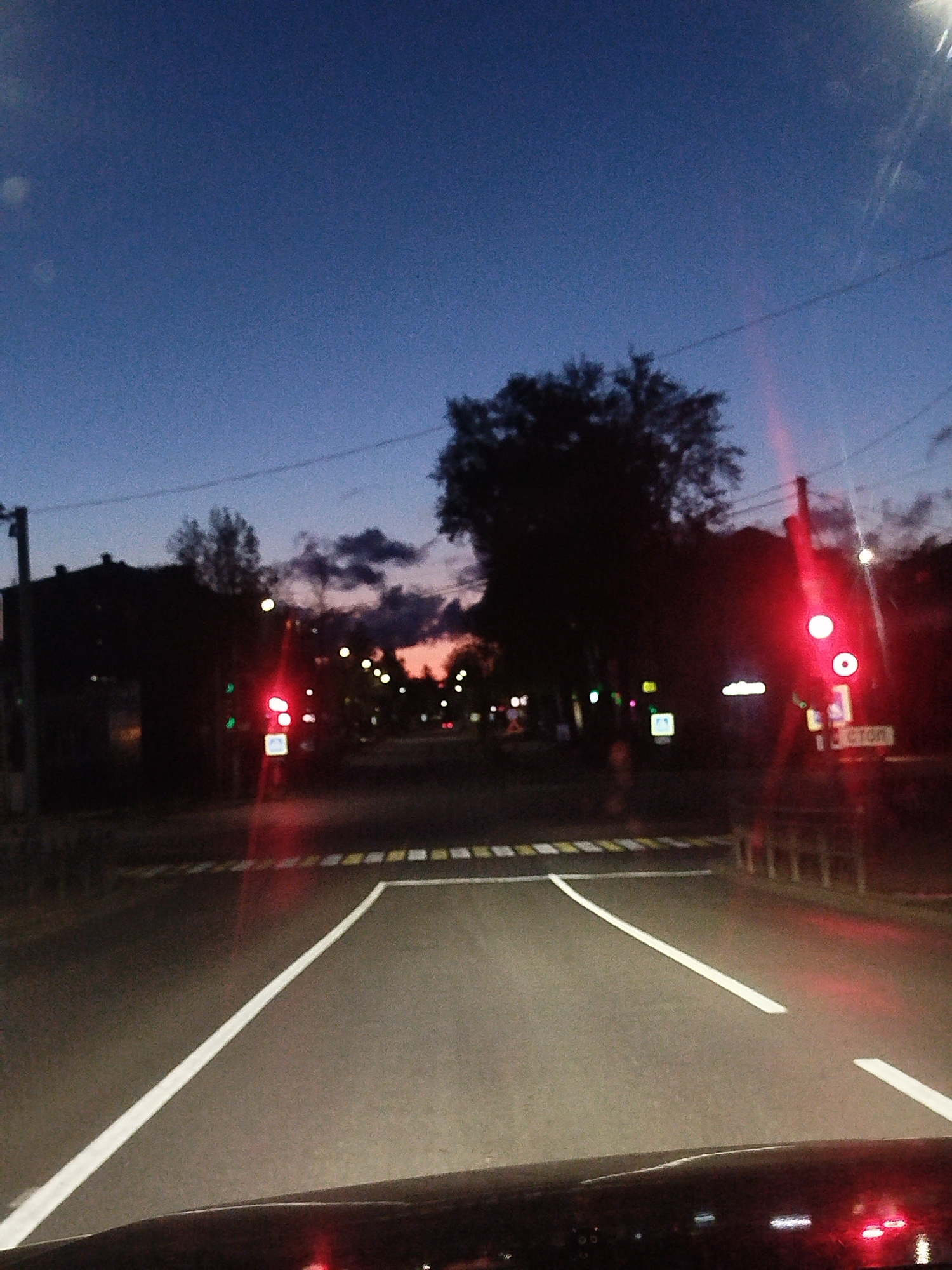 A street scene in Chaikovsky, Russia at twilight. The view is from the driver's seat of a car, stopped at a stop line at an intersection. The sky is a deep blue with some clouds visible near the horizon. The road ahead is asphalt, marked with white lines. A pedestrian crossing is visible ahead with alternating yellow and white stripes. A traffic light on the right shows a red signal. The traffic light has a white sign below it with the word "СТОП." Other buildings and trees are visible in the background with street lights illuminated. A utility pole is visible on the left.