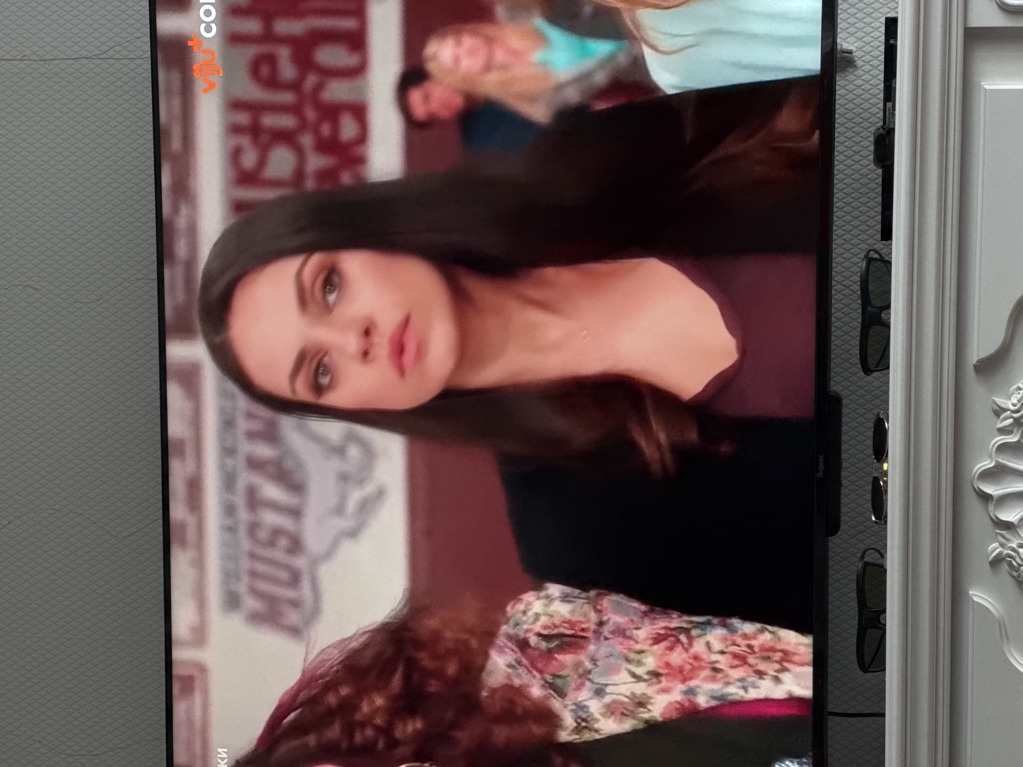 The image is a medium shot of actress Mila Kunis on a television screen. She is wearing a dark blazer over a burgundy top, and her dark hair is down. She is looking off to the side with a serious expression. In the background, there are other people visible, out of focus, and a sign that reads "MUSTANG". The television is placed on a white mantelpiece with two pairs of sunglasses on it. The overall setting appears to be indoors, likely a living room. The specific location in Oskemen, Kazakhstan cannot be determined from the image.