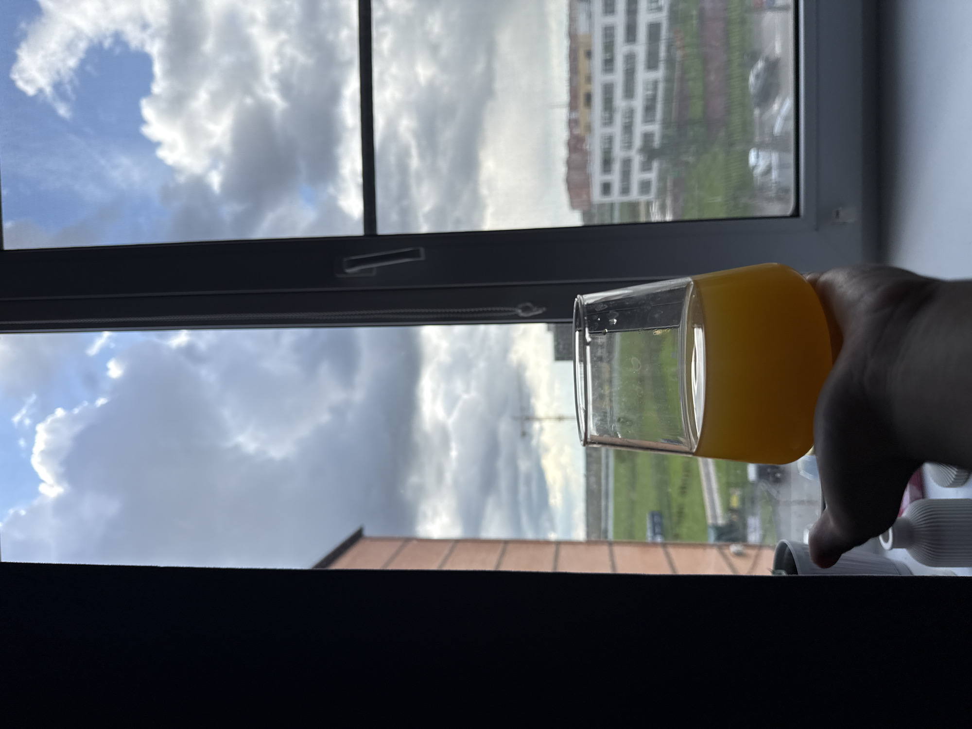 A glass of orange juice is held in front of an open window. Outside the window, the sky is cloudy. In the background, a building is visible.