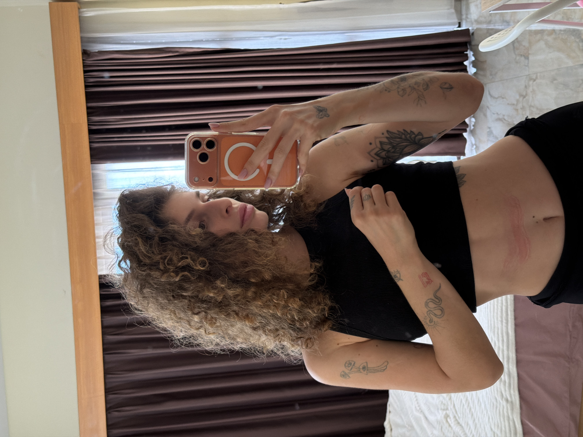 A mirror selfie captures a young woman with voluminous, curly brown hair, standing indoors, likely in a residence in North Kuta, Indonesia, during daylight hours. She is holding a smartphone, which appears to be an iPhone with multiple cameras and a clear case featuring an orange MagSafe-compatible ring, positioned to take the picture. Her expression is neutral as she looks towards the phone.
She is wearing a black ribbed crop top, exposing her midriff, where a distinct reddish scratch or mark is visible on her left side (viewer's right). Her arms and hands are extensively tattooed, including a prominent mandala on her upper right arm (viewer's left), a small butterfly on her right forearm (viewer's left), a snake design, an eye symbol, and other abstract patterns on her lower arms. A small crown or symbol is tattooed on one of her fingers.
The background of the mirror's reflection includes a light wooden frame on the left. Behind her, there are dark brown horizontal blinds or curtains, with a window above them allowing natural light to enter. To the right of the woman, a light-colored, possibly marble-tiled wall is visible, along with a white, indistinct object, potentially a hanger. In the lower right corner of the reflection, a bed is visible with a white textured blanket covering a pink or purple sheet. No discernible text is visible in the image.