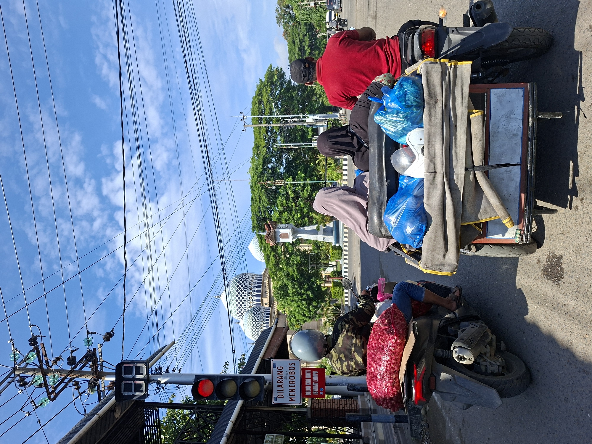 A sunny day in Lhokseumawe, Indonesia is depicted in this image, with a clear blue sky dotted with white clouds. The scene is dominated by a bustling street with several vehicles and pedestrians. In the foreground, a motorcycle is equipped with a sidecar, carrying passengers and goods. The motorcycle itself is laden with a large sack of red onions. Behind it, another motorcycle carries a rider wearing a camouflage jacket and jeans, with a red sack of onions secured behind them. The sidecar contains three passengers: two women wearing hijabs, one in light purple and the other in black, and a man in a red shirt and black cap. Blue plastic bags and a white helmet are also visible in the sidecar.
Traffic signals and electrical wires crisscross the sky, indicating an urban setting. A prominent mosque with distinctive patterned domes serves as a backdrop, its white and green architecture standing out against the greenery of trees. A clock tower with a traditional design is also visible near the mosque. A traffic light displaying a red signal suggests the vehicles are stopped. A sign on a pole reads "DILARANG MENEROBOS BELOK KIRI IKUTI ISYARAT LAMPU," which translates to "DO NOT PROCEED, TURN LEFT FOLLOW THE LIGHT SIGNAL." The road is asphalt, with some wear and tear evident, including what appears to be a spill on the surface. The overall atmosphere is that of a typical day in an Indonesian city, with daily life unfolding on the streets.