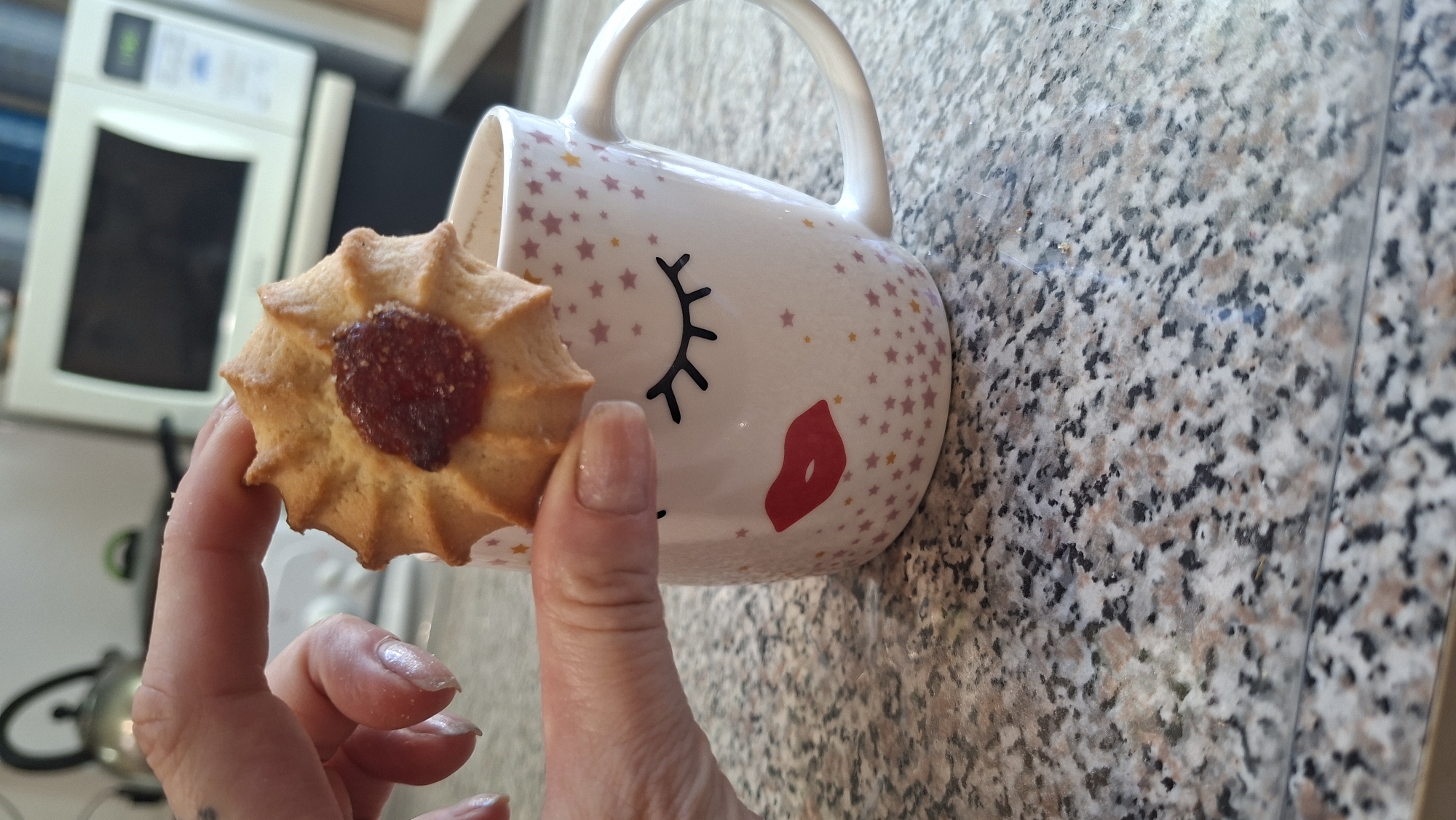 A close-up shot shows a person's hand holding a jam-filled cookie over a mug. The mug is white with a design of closed eyes and red lips, and is decorated with pink and yellow stars. The background shows a microwave oven and kitchen counter. This image was taken in Myadzyel, Belarus.