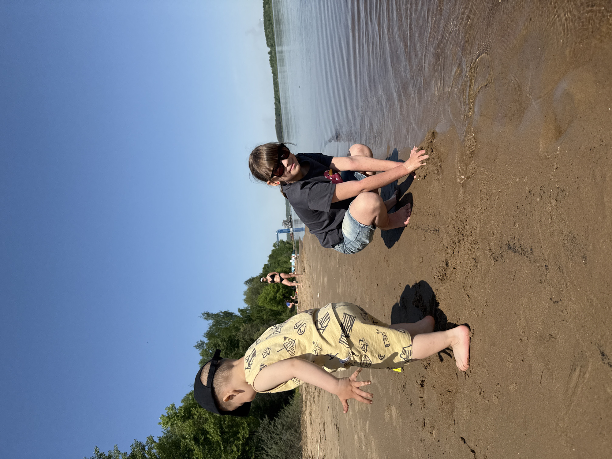 Two children are on a sandy beach near water under a clear blue sky. An older child wearing sunglasses, a dark shirt, and jean shorts is squatting at the water's edge, touching the sand. A younger child wearing a black cap and yellow patterned shorts is crawling on the sand. In the background, there is a body of water, trees, and other people on the beach. The location is Unknown, Unknown.
