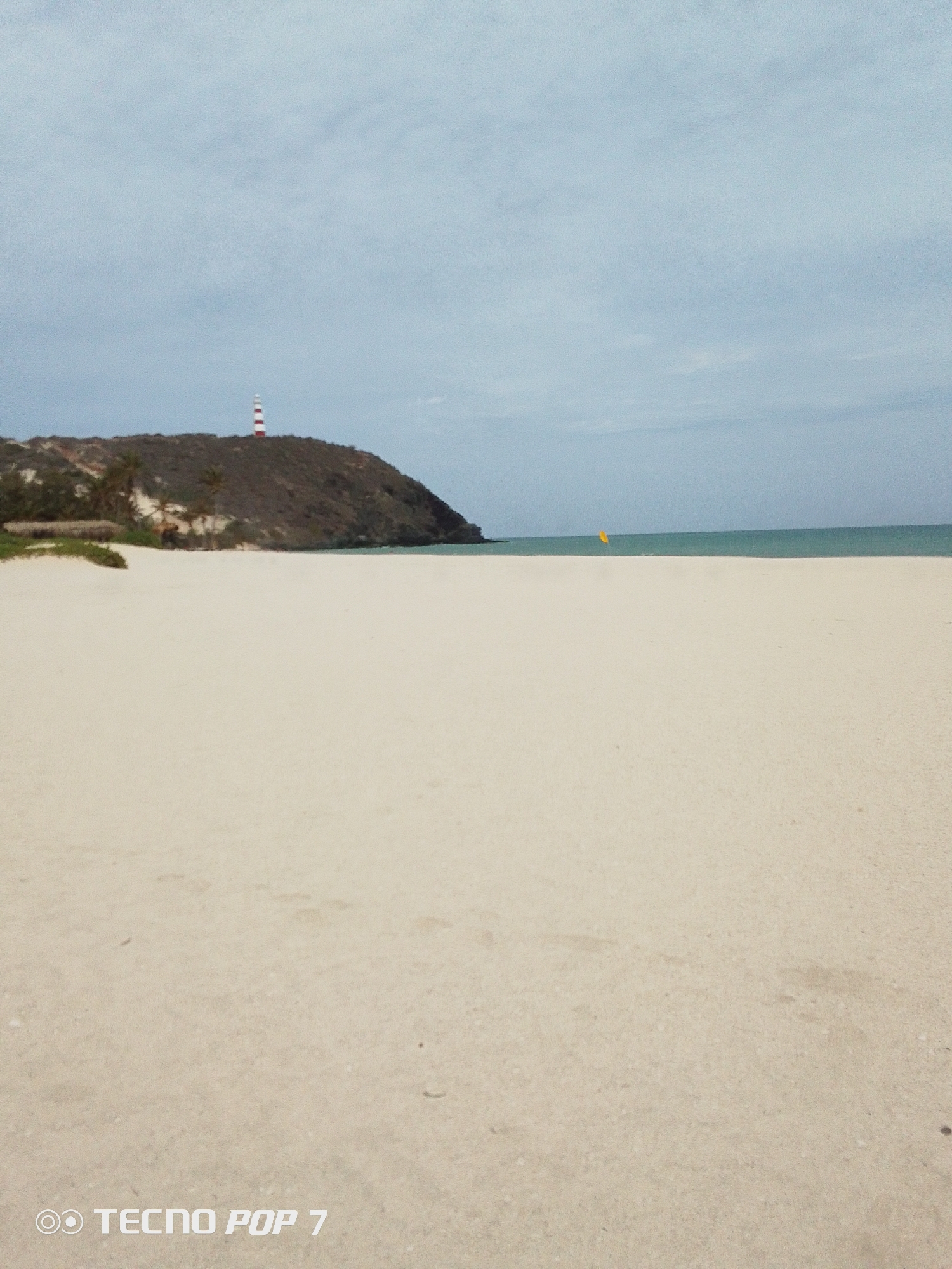 The scene is a beach in Pedro González, Venezuela, with a prominent sandy shoreline in the foreground. Behind the beach is a small, brown, vegetated hill. A red-and-white striped lighthouse stands atop the hill. In the distance, the ocean meets the sky, under an overcast sky. A yellow object is on the ocean line. The image was captured on a TECNO POP 7 device.