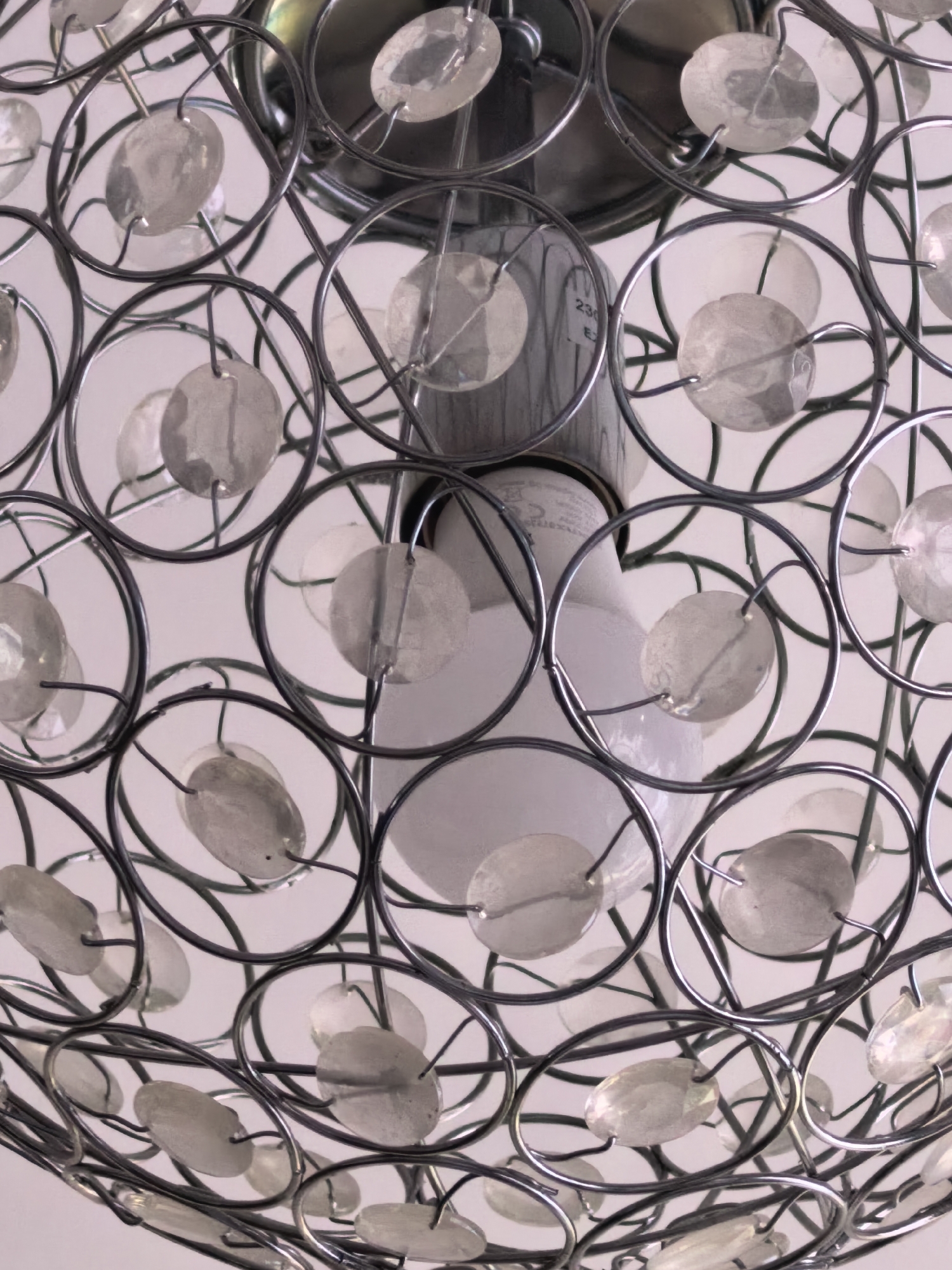 A close-up, eye-level shot focuses on a light fixture. The fixture is comprised of a series of silver-colored metal circles interconnected to form a cage-like structure. Within each circle, a small, opaque disc is attached by thin metal wires. The light bulb is located in the center of the fixture. Part of the light fixture's mounting hardware, also in silver-colored metal, is visible at the top of the frame. The background is a soft, off-white color, likely a ceiling. No location is provided.