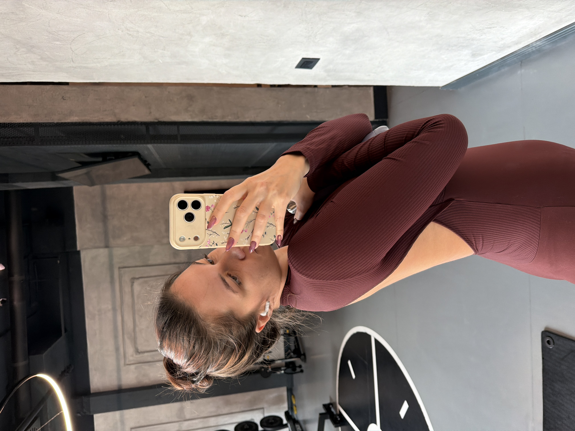The image, when properly oriented, shows a woman taking a mirror selfie or a selfie using her phone's rear camera in what appears to be a modern gym or fitness studio. She is seen from the upper torso, holding a light-colored smartphone up, looking intently at its screen.
The woman has light brown hair, possibly tied back in a bun, and wears a form-fitting, long-sleeved athletic top in a dark reddish-brown (maroon or burgundy) color, featuring a ribbed texture and a high neckline. Small white wireless earbuds are visible in her ears. Her nails are painted with a light, possibly purple or pink, polish, and she has delicate rings on her fingers.
The smartphone is a beige or cream color with a triple camera lens array visible on its back. The phone case has a decorative floral pattern featuring pink or purple flowers and green leaves on dark branches.
The background indicates an indoor setting with a contemporary design. A light grey, possibly textured, wall or ceiling is visible at the top, transitioning to a darker grey or brown section below it, which could be a beam or decorative panel. In the lower left portion of the background, black exercise weights or plates are partially visible on a rack. To the woman's right, a large circular black and white graphic, resembling a target or clock face, is mounted on a light grey wall. A reflective surface or light fixture creates a distinct ring of light in the bottom left, with other dark structural elements and equipment visible. The setting is consistent with a fitness studio, located in Belgrade, Serbia.