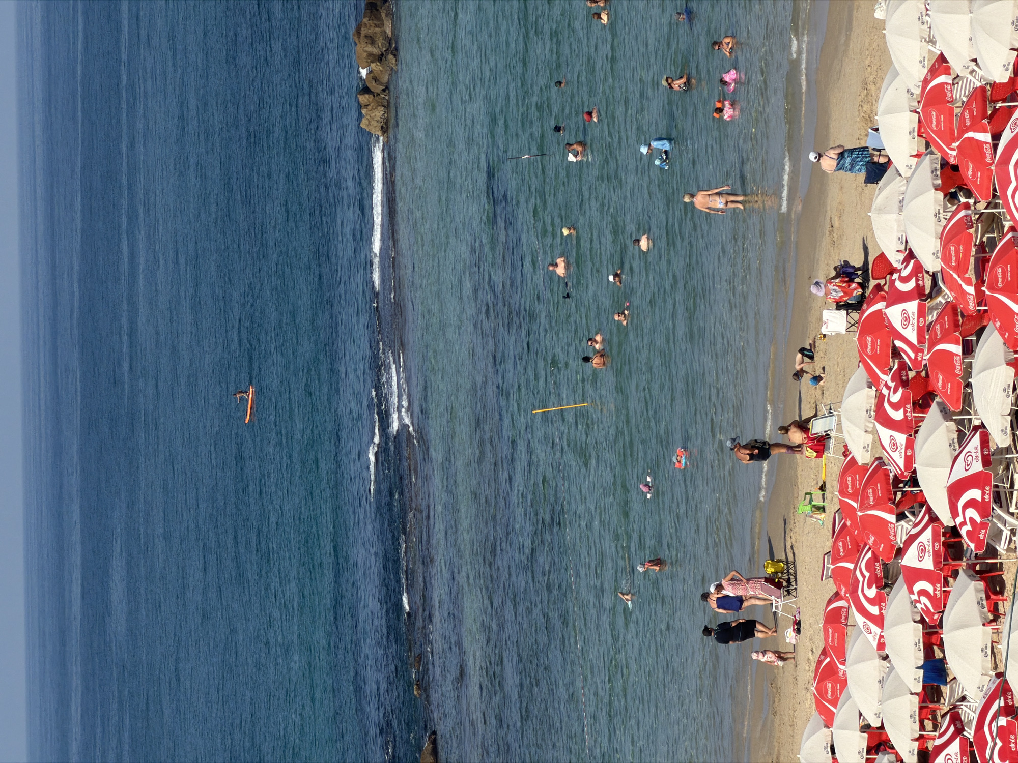 The image shows a crowded beach scene on a sunny day in Bat Yam, Israel. The foreground is filled with numerous red and white beach umbrellas, many of which are branded with "Coca-Cola" and "Corona". Several beach chairs are visible beneath the umbrellas.
The middle ground depicts people swimming and wading in the shallow, clear turquoise water. A group of individuals are clustered closer to the shore, while others are dispersed further out. Some are floating on inflatables or using small boards. Near the shore, a few people are standing on the sand, and a child is playing.
In the background, the vast blue expanse of the Mediterranean Sea stretches to the horizon. A single person is visible on an orange paddleboard in the distance. The water has gentle waves breaking near a rocky outcrop on the right side of the frame. The sky is a hazy blue.