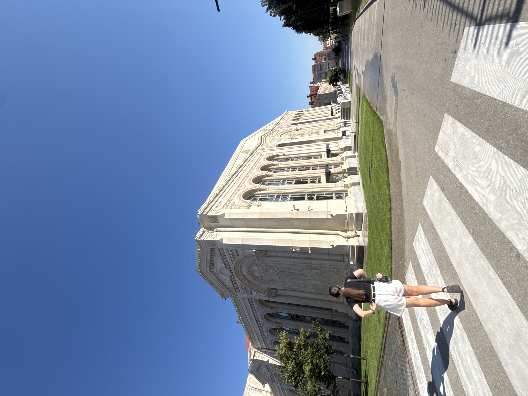 A person is walking across a pedestrian crossing in front of a large, ornate building with a clear blue sky above. The building appears to be made of light-colored stone and features arched windows and decorative carvings. The person is wearing a black t-shirt, white shorts, and black sandals. They are facing away from the camera and have their hands on their hips. There is a green lawn in front of the building, and a tree is visible to the left. The pedestrian crossing is marked with white stripes on an asphalt road. The scene appears to be outdoors on a sunny day. The building is the St. Volodymyr's Cathedral in Sevastopol, Ukraine.